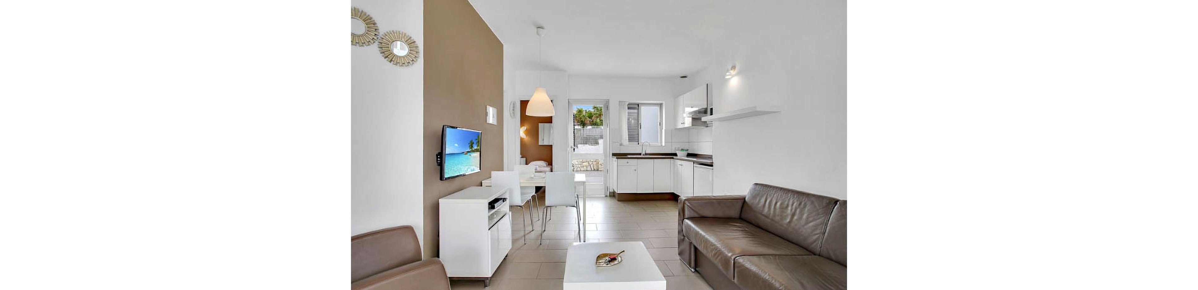 A contemporary living space features a brown leather sofa, wall-mounted TV, and a white dining set. The open-plan layout connects the living area to a minimalist kitchen with white cabinetry and stainless steel appliances. Neutral tones and natural light create a bright, inviting atmosphere, with decorative mirrors and a pendant lamp adding visual interest.