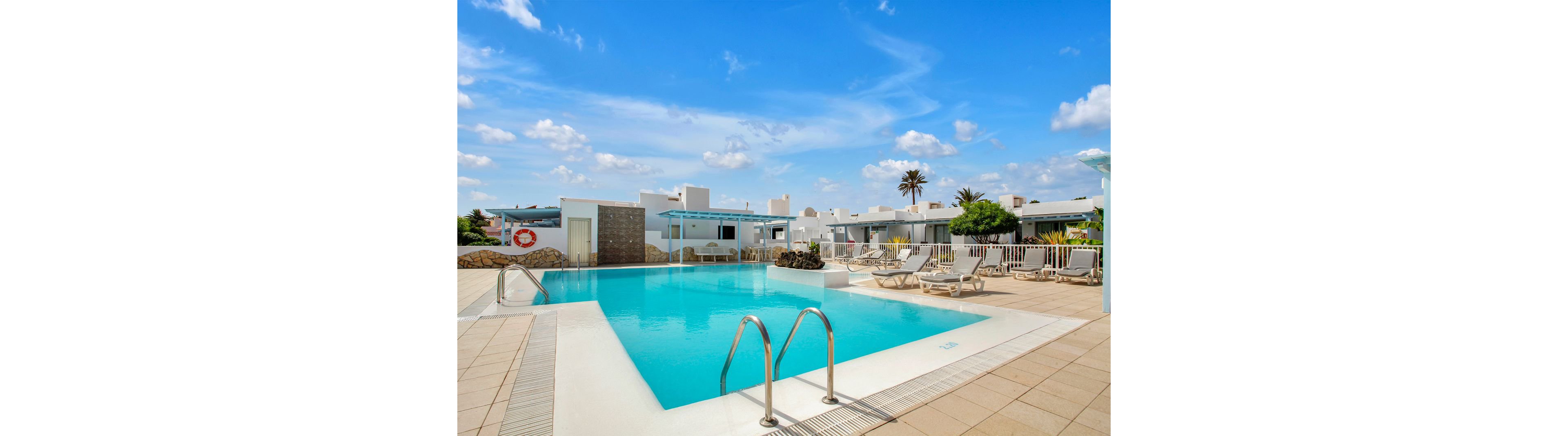 A large, rectangular swimming pool is the central feature of a modern resort courtyard. White lounge chairs and umbrellas line the poolside, with white buildings and palm trees in the background. The setting is bright and sunny, with a clear blue sky and scattered clouds. The mood is relaxed and inviting, ideal for vacation or leisure. A red and white life preserver is visible on the wall.