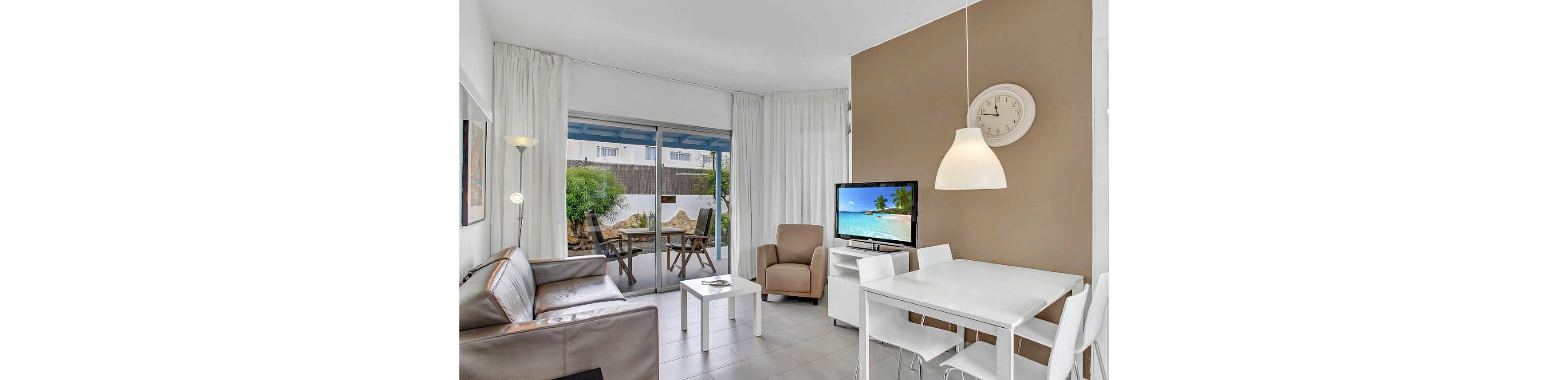 A contemporary living room features a brown sofa, armchair, and a small white dining table set for two. The space is well-lit with natural light from large sliding glass doors leading to an outdoor patio. A wall clock shows the time as 10:10, and a television displays a tropical beach scene. The room is decorated in neutral tones with white curtains and minimalist furnishings.
