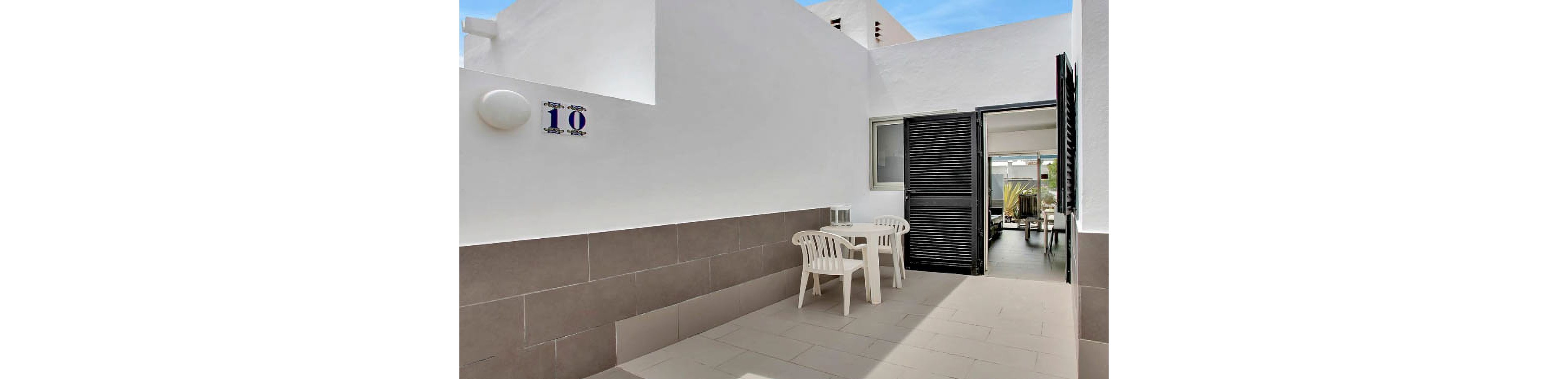 A clean, modern outdoor patio features two white plastic chairs and a small table with a wine bottle and glasses. The setting is adjacent to a white building with a black door and open shutters, leading into a bright interior. The number '10' is clearly visible on the wall, and the overall palette is neutral with white and gray tones. The scene suggests a relaxed, inviting atmosphere for outdoor dining or leisure.