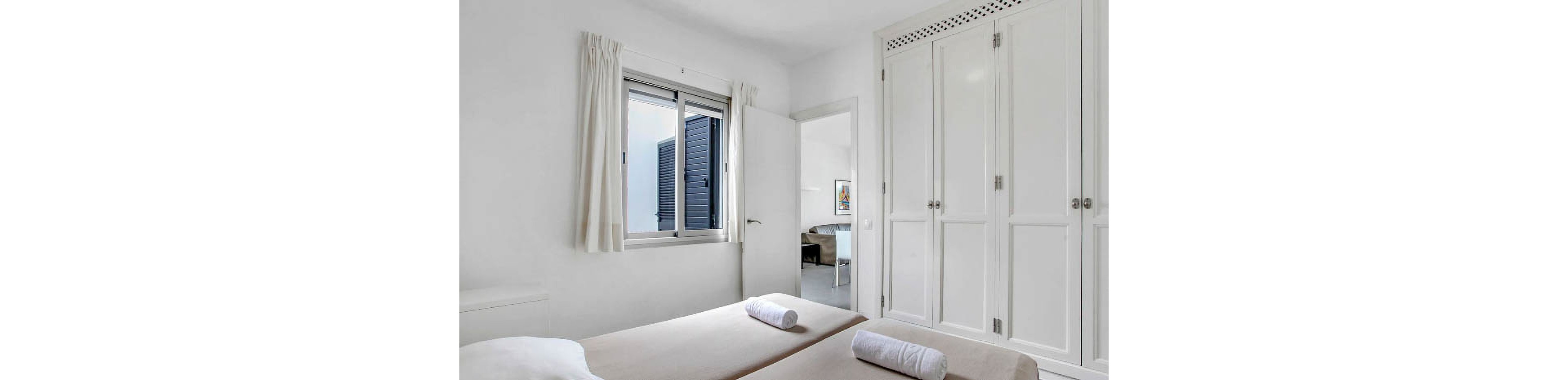 A clean, modern bedroom features two twin beds with beige linens and rolled white towels. The space is illuminated by natural light from a window with white curtains, and includes a large built-in white wardrobe. The open door reveals a glimpse into an adjacent room with contemporary furnishings. The overall palette is neutral, creating a calm and inviting atmosphere.