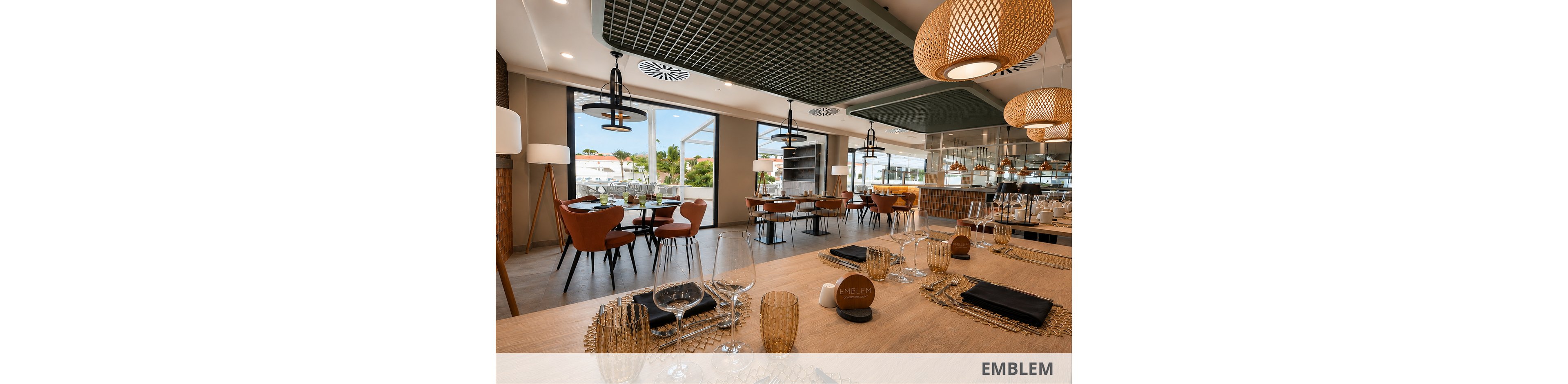 A contemporary restaurant dining room features neatly arranged tables set with glassware, plates, and dark cloth napkins. Warm wooden tones, woven pendant lights, and tripod floor lamps create a cozy yet upscale atmosphere. Large windows reveal an outdoor view with greenery and buildings in the distance. Reflections in the mirrored wall enhance the sense of space and highlight the stylish decor.