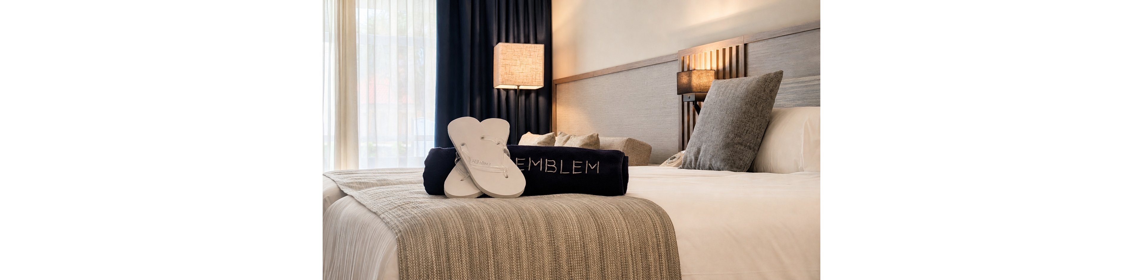 A neatly made hotel bed features a folded dark towel and a pair of white flip-flops placed on top. The word "EMBLEM" is clearly visible on the towel, suggesting a named property or collection. Warm bedside lamps and a wooden headboard add a soft, inviting glow to the room. Sheer curtains allow natural light to filter in, complementing the neutral beige and gray color palette.