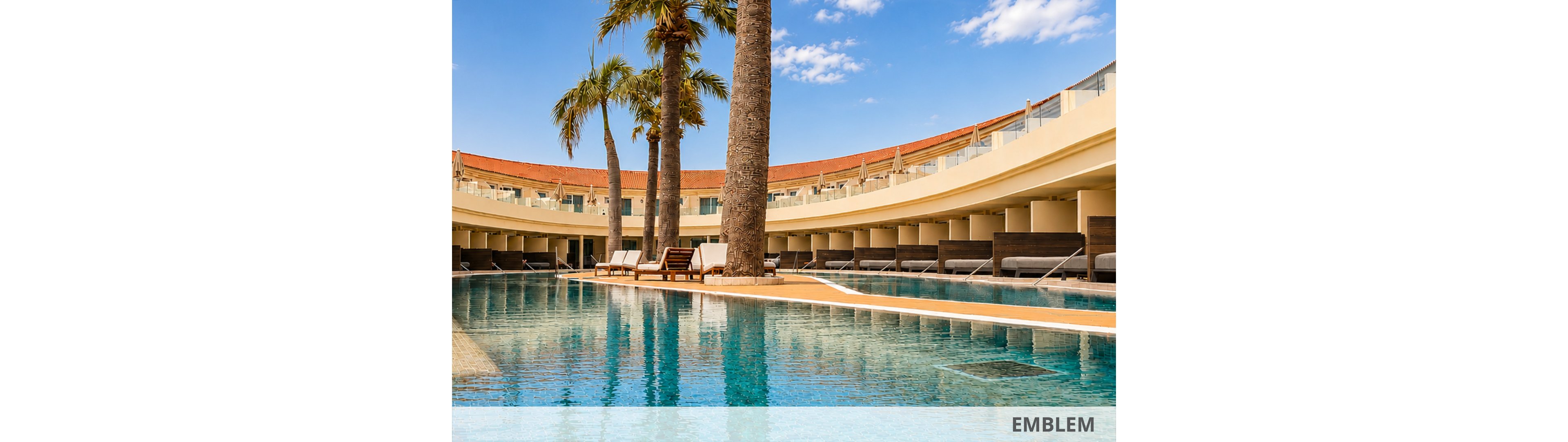 A large, clear blue swimming pool sits at the center of a modern resort courtyard. Tall palm trees rise from islands within the pool, adding a tropical touch to the contemporary architecture. Curved rows of sun loungers and shaded daybeds line the water’s edge beneath a sweeping terrace. The bright daylight and blue sky create a warm, inviting vacation atmosphere.