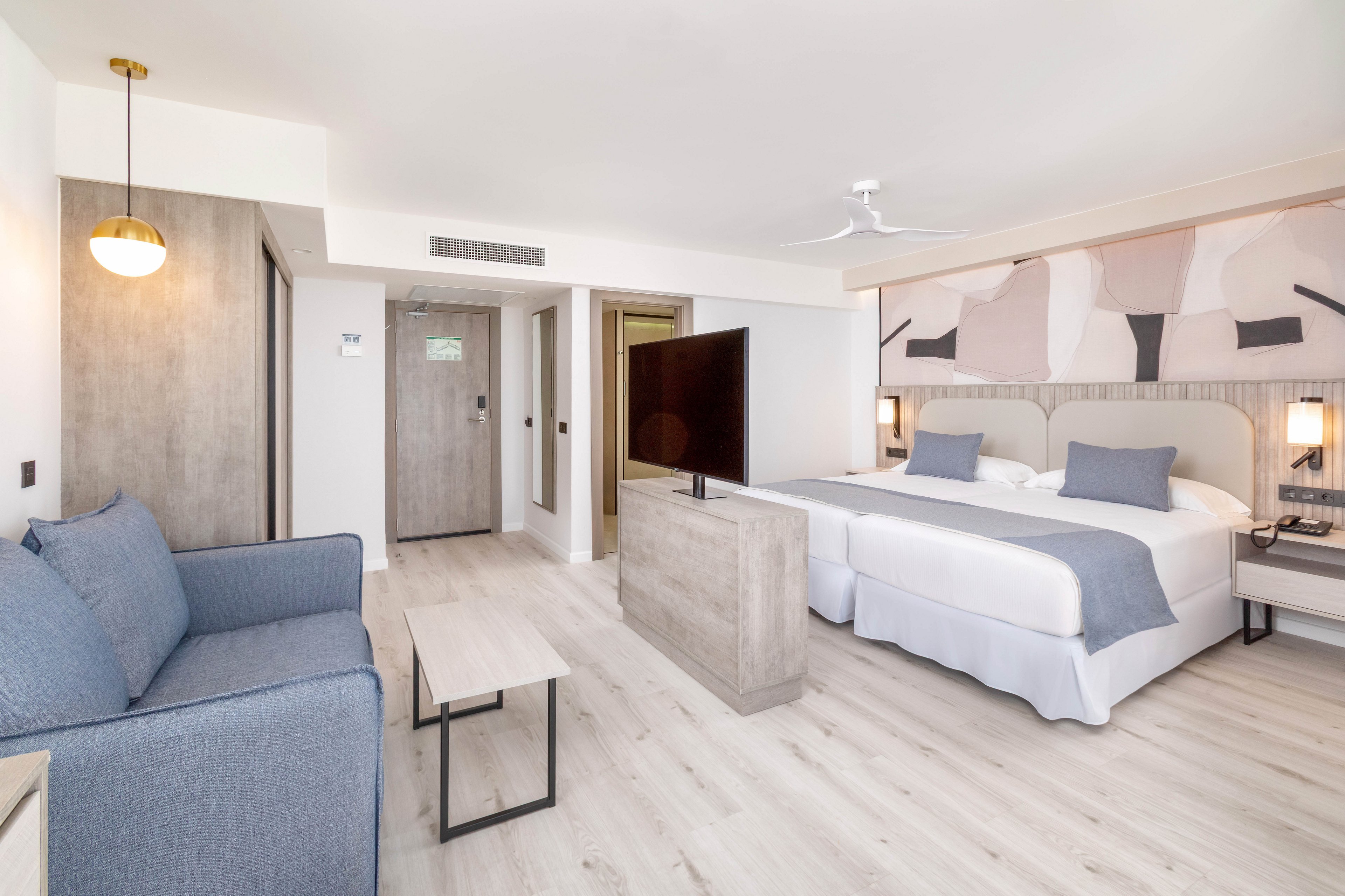 A spacious hotel room features two twin beds with gray accent pillows and throws, a blue sofa, and a flat-screen TV on a central stand. The setting is bright and airy, with light wood flooring and abstract wall art above the beds. The room includes a minimalist coffee table, pendant lighting, and a ceiling fan, creating a contemporary and comfortable atmosphere.