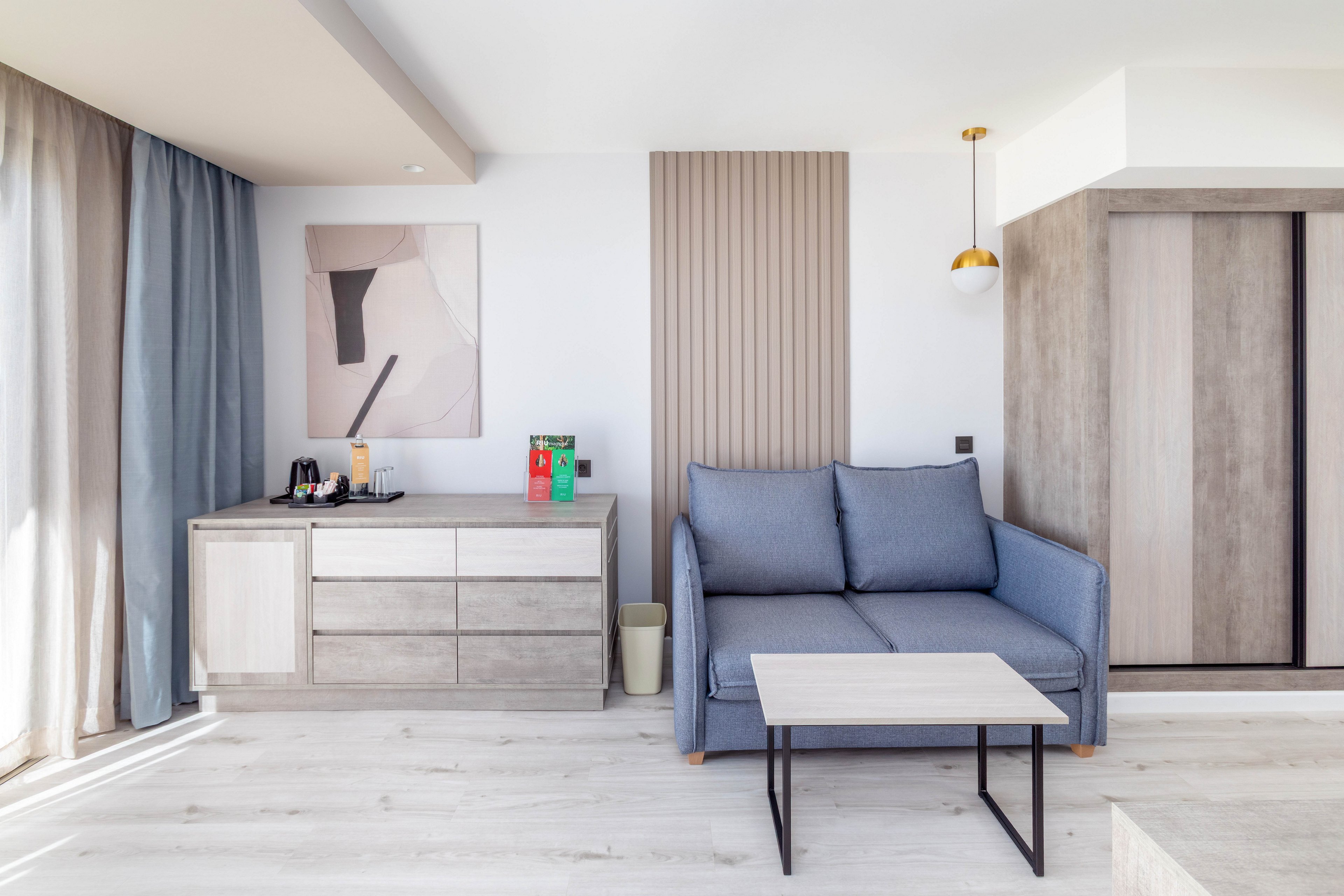 A bright, contemporary hotel room features a blue sofa and a light wood coffee table. To the left, a refreshment station with a kettle and cups sits atop a cabinet, with a colorful box displaying the number '20'. The room is decorated in neutral tones with minimalist artwork and soft natural light from large windows. No people are present in the scene.