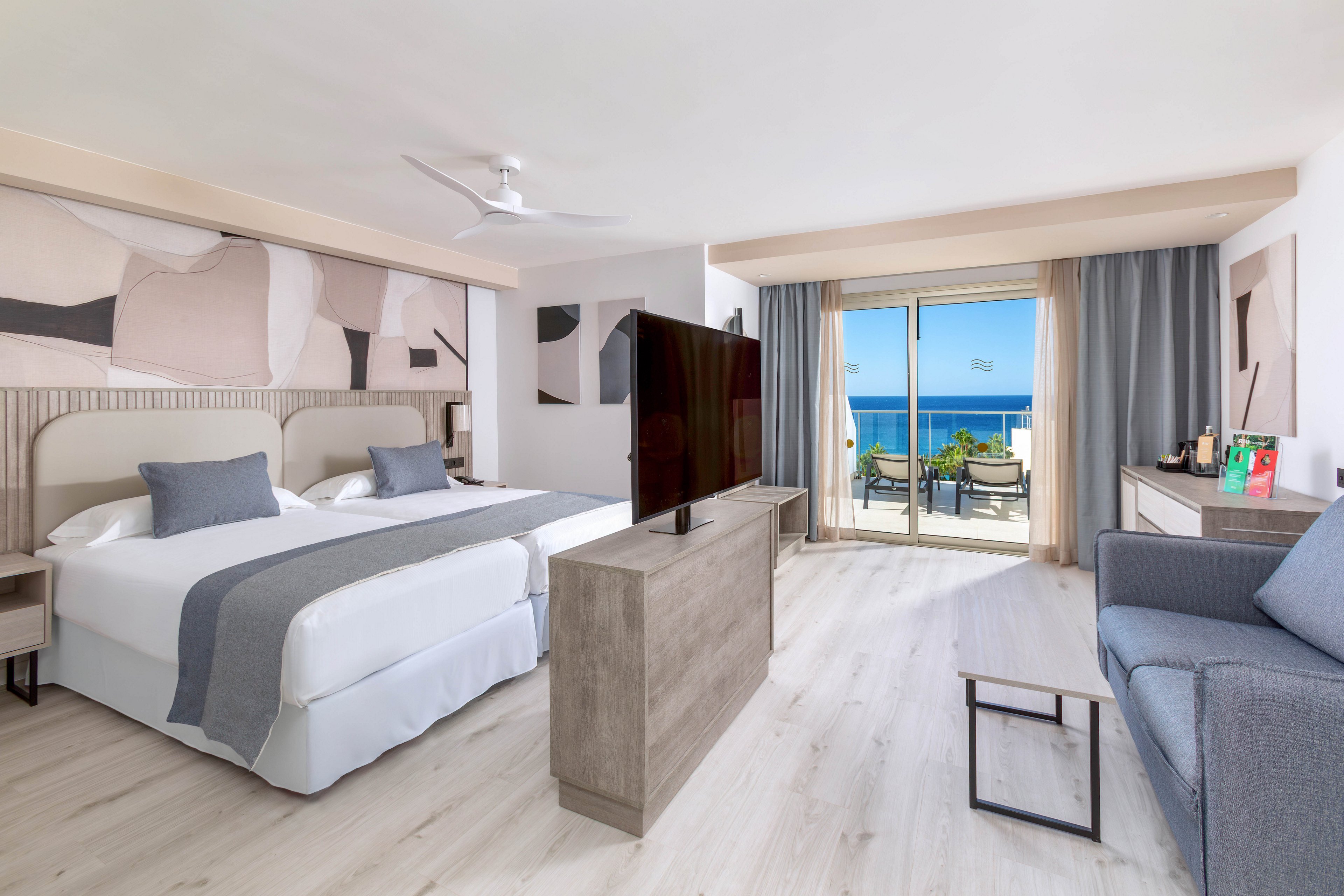 A spacious hotel room features two large beds with gray accents, a flat-screen TV, and a comfortable seating area. Abstract wall art decorates the space, and a sliding glass door opens to a balcony overlooking the blue ocean. The room is bright and airy, with light wood flooring and contemporary furnishings.