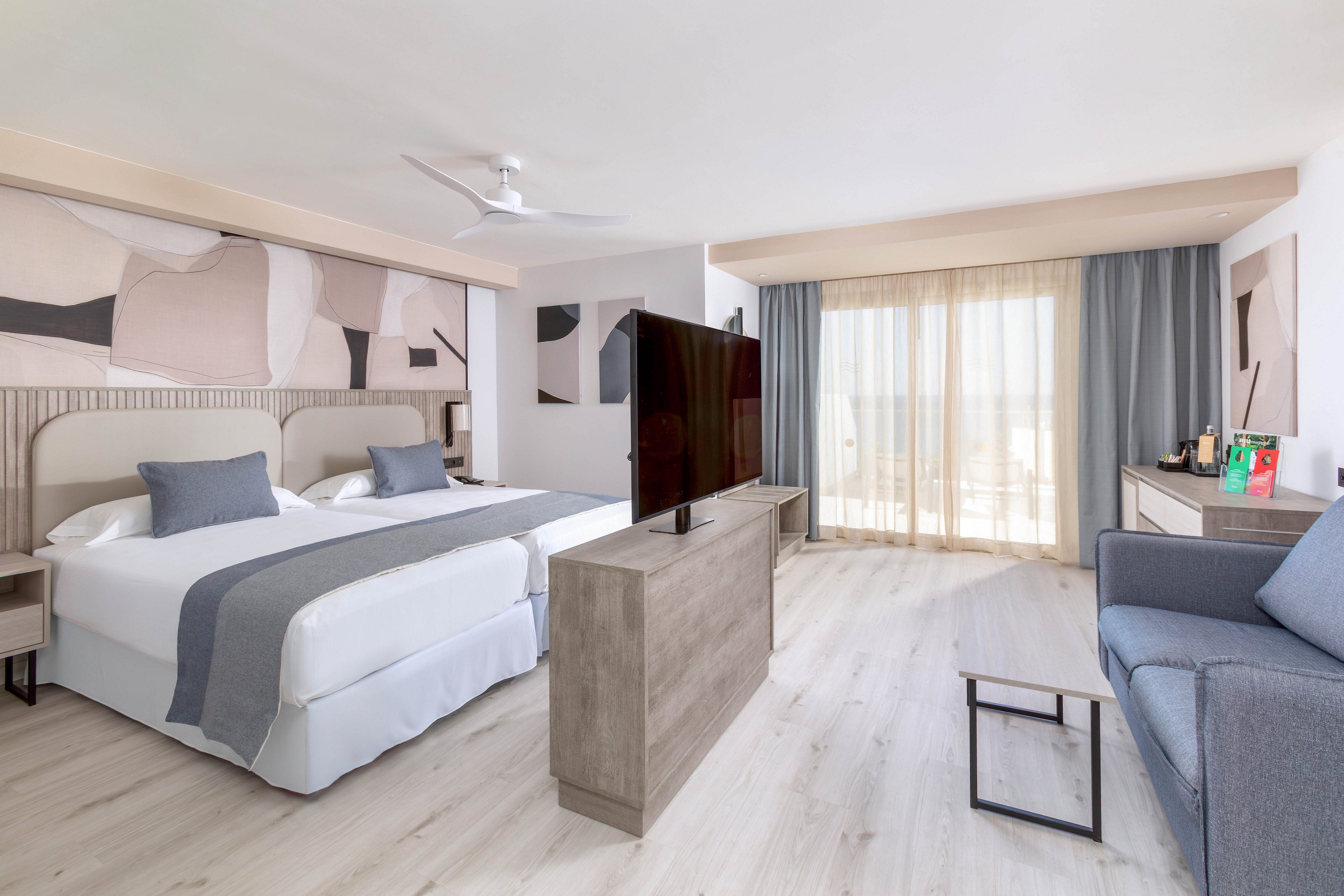 A spacious hotel room features two twin beds with gray accents, a large flat-screen TV, and a comfortable seating area. The room is decorated in a contemporary style with abstract wall art and light wood flooring. Natural light streams in through sheer curtains covering a balcony door, creating a bright and airy atmosphere. A desk with amenities is visible, enhancing the functional appeal of the space.