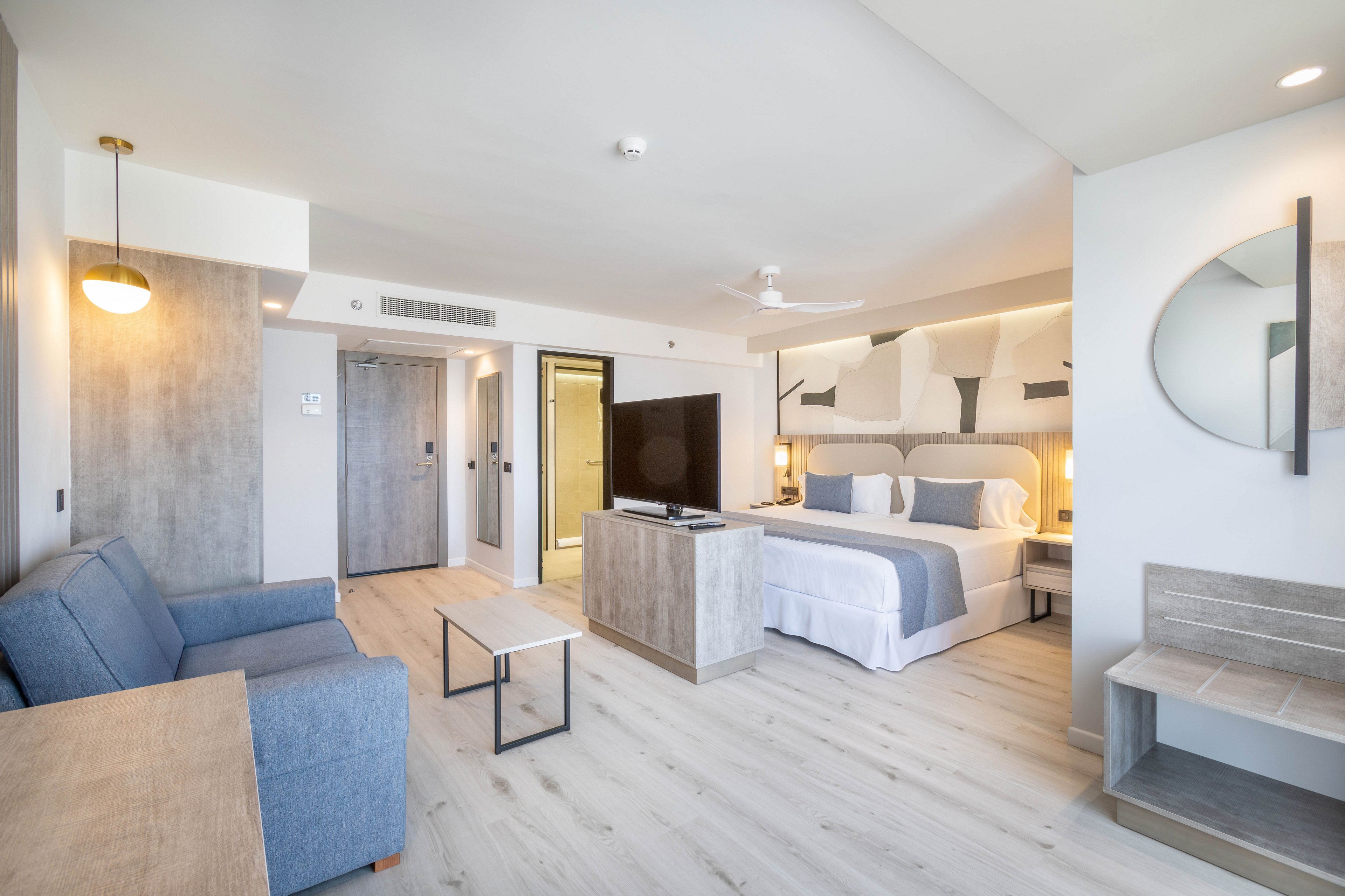 A spacious, contemporary hotel room features a king-sized bed with crisp white linens and accent pillows. The suite includes a blue sofa, a flat-screen television, and minimalist furnishings in neutral tones. Light wood flooring and soft lighting create a calm, inviting atmosphere. The room is well-lit with natural light from large windows, and a round mirror decorates one wall.