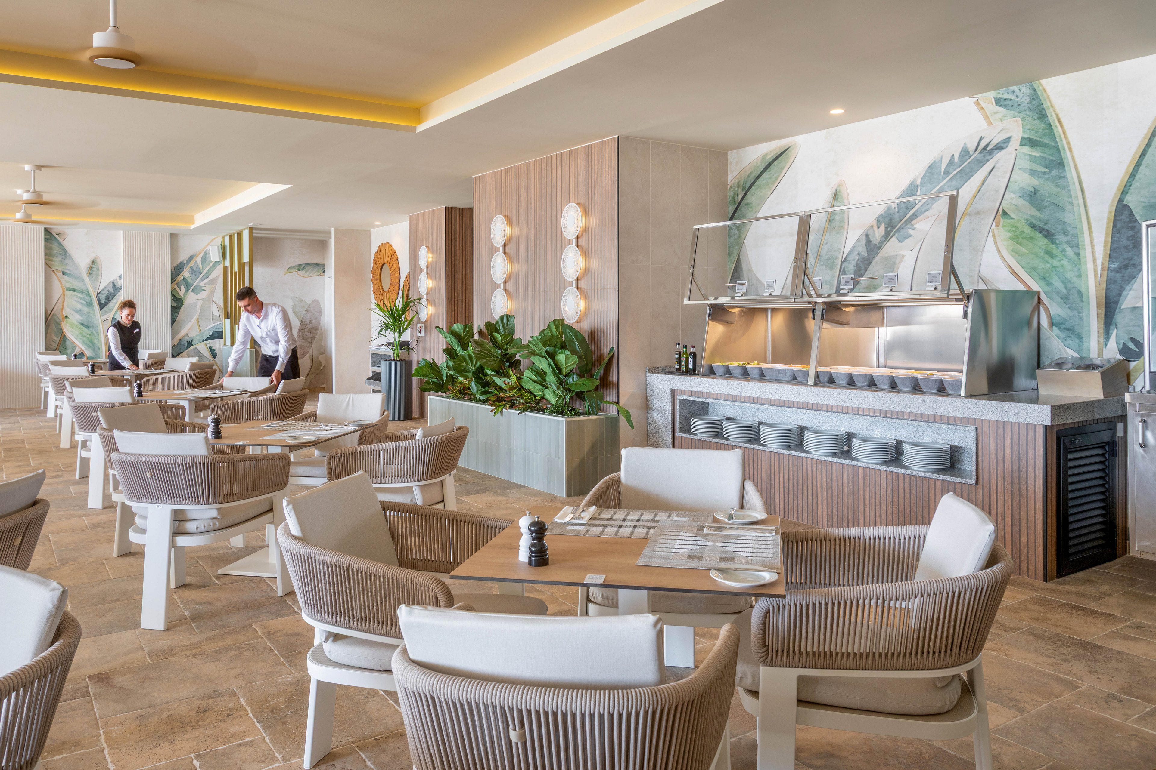 The image shows a contemporary restaurant dining area featuring neutral-toned furniture and tropical leaf murals on the walls. Two staff members are visible, one setting a table and another walking, suggesting a welcoming and service-oriented atmosphere. The space is well-lit with natural and artificial lighting, and includes a buffet counter with stacked plates. The mood is relaxed and inviting, with a clean and modern aesthetic.