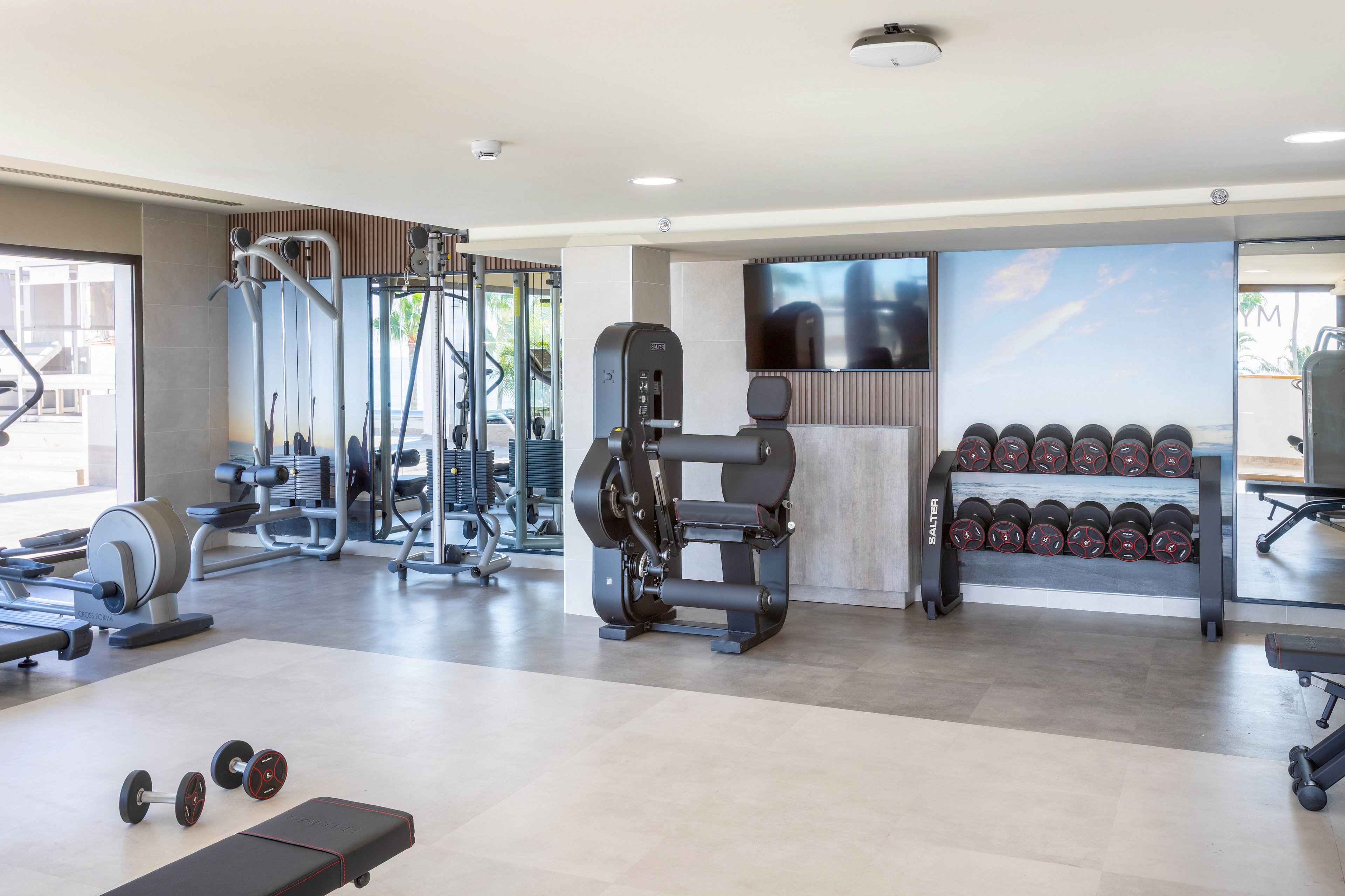 A spacious, well-lit gym features a range of exercise machines, free weights, and benches. The setting is clean and contemporary, with large windows allowing natural light to fill the room. Dumbbells, weight racks, and cardio machines are neatly organized, and a wall-mounted television is visible. The color palette is neutral with gray and white tones, creating a calm and motivating atmosphere.