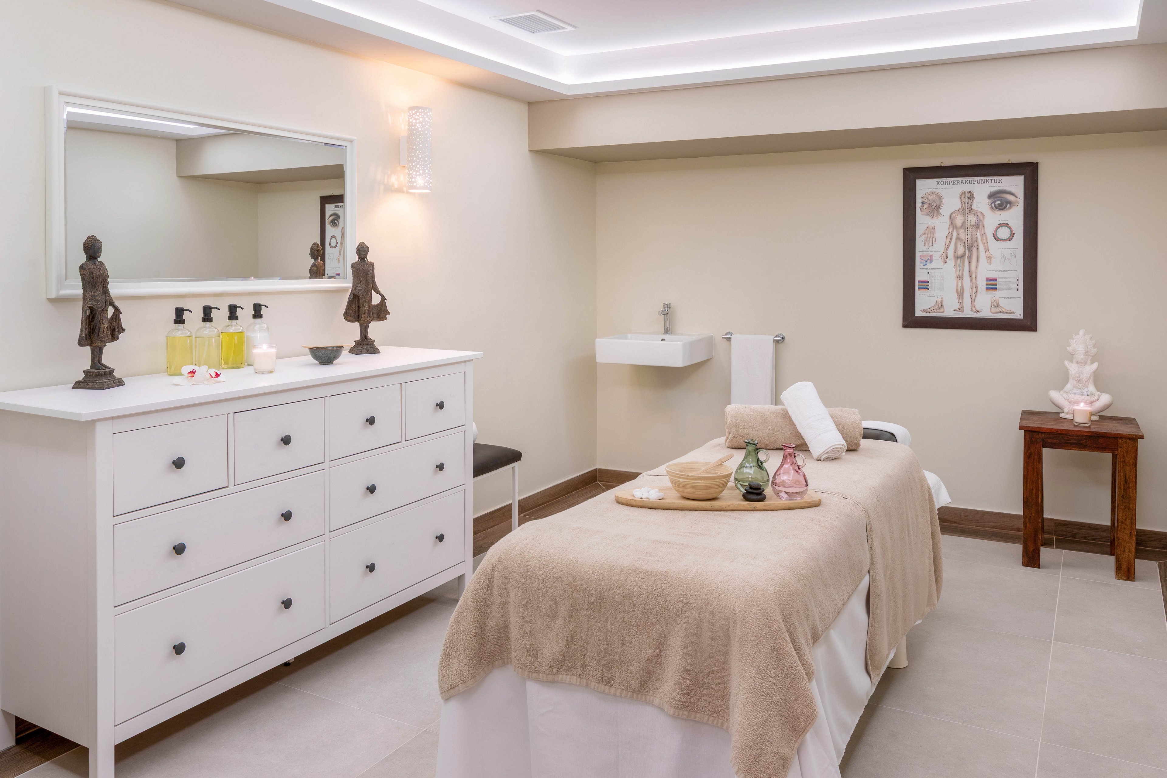 A contemporary spa room features a massage table covered with beige linens and neatly arranged towels. The setting includes a white dresser with skincare bottles, decorative statues, and a wall chart displaying anatomical diagrams. Soft lighting and neutral tones create a calm, relaxing atmosphere, with a sink and small table adding functional elements.