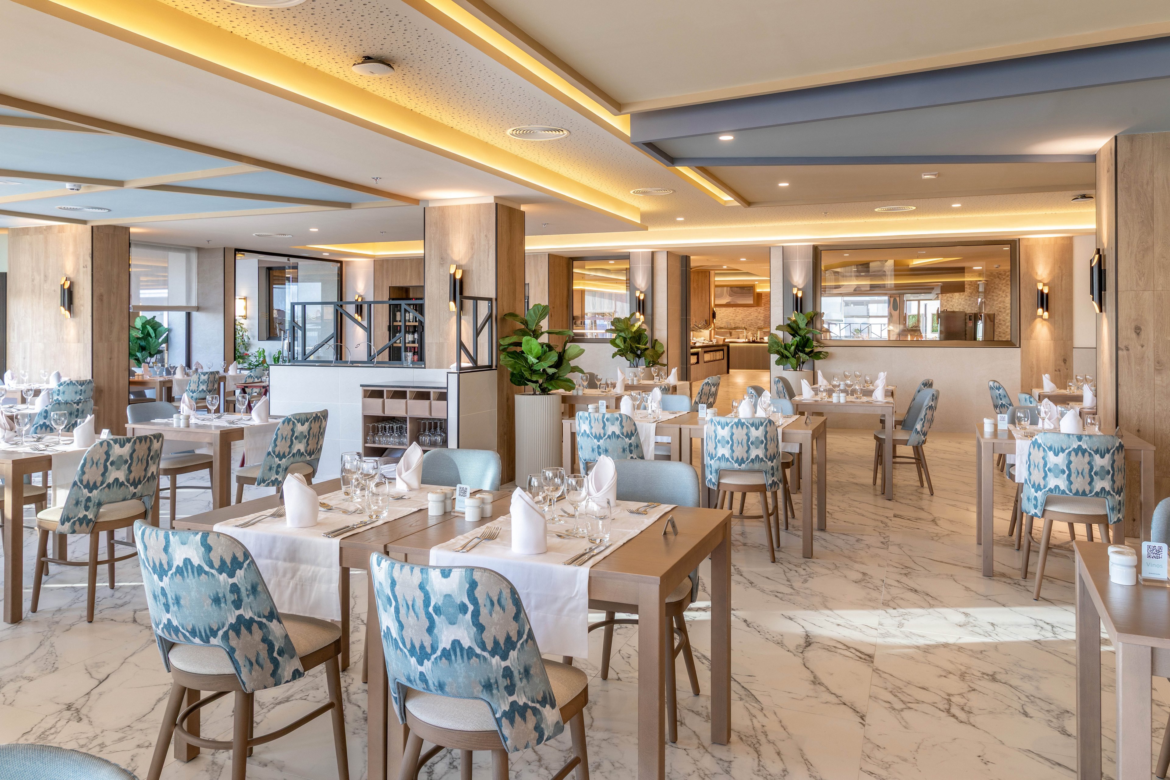 The image shows a spacious, contemporary restaurant dining area featuring patterned blue and white chairs and wooden tables. The setting is bright with natural light, marble flooring, and neatly arranged tableware, including glasses and napkins. The mood is inviting and upscale, with a neutral palette accented by blue tones and greenery. No visible text or numeric values are present in the scene.