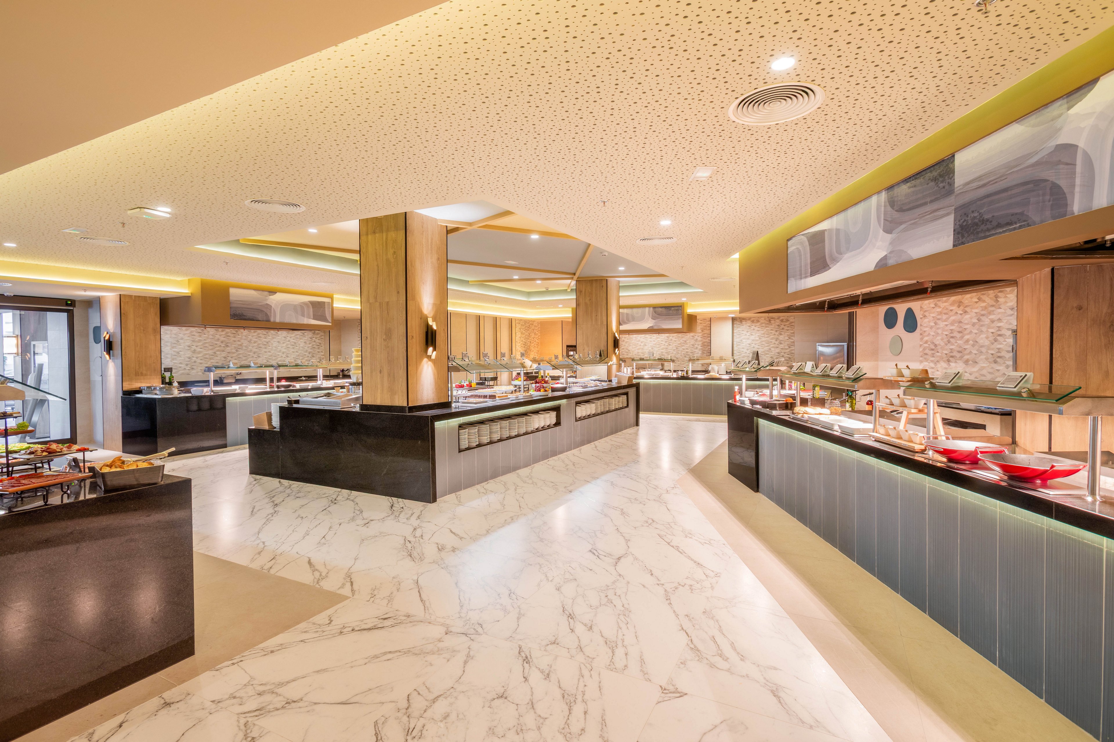 The image shows a spacious buffet area in a contemporary hotel or restaurant. The setting features sleek counters, marble floors, and warm ambient lighting, with food stations arranged for self-service. The atmosphere is clean and inviting, with neutral tones and minimalist decor. No visible people or text are present in the scene.