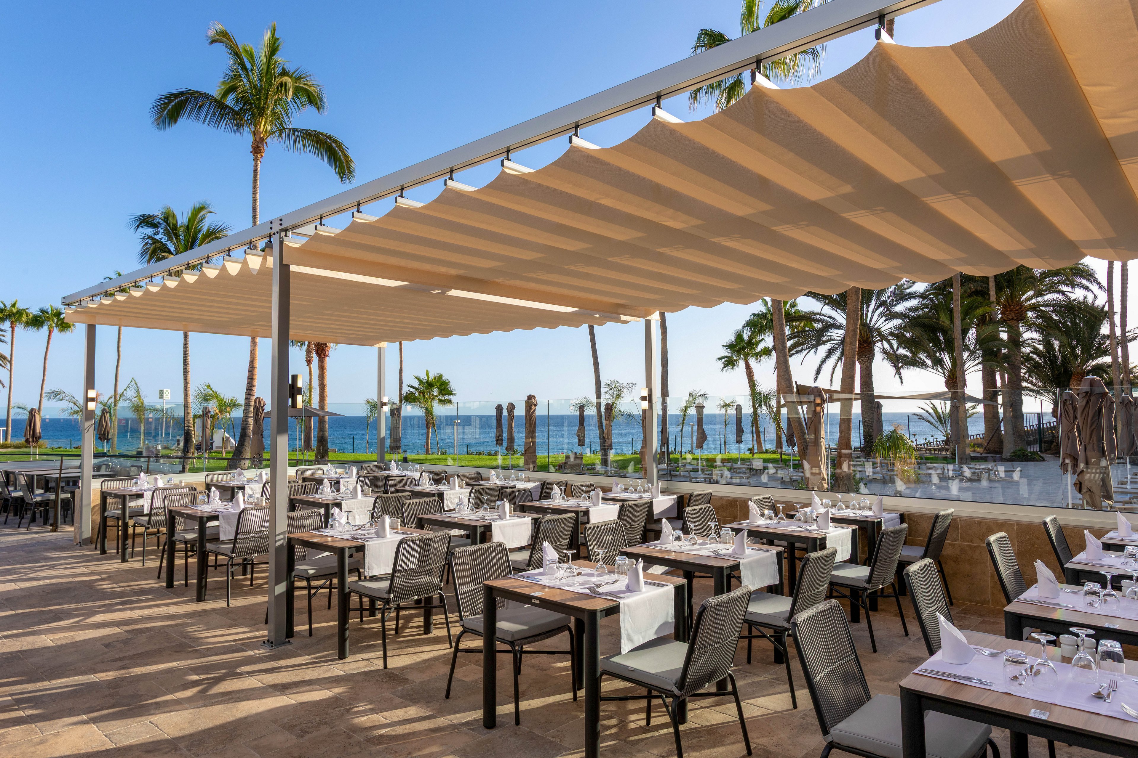 The image shows a spacious outdoor restaurant with neatly arranged tables and chairs under a large beige canopy. The setting overlooks a scenic ocean view, surrounded by palm trees and clear blue skies. The tables are set with white linens and glassware, creating a relaxed yet elegant atmosphere. No visible text or numeric values are present in the scene.