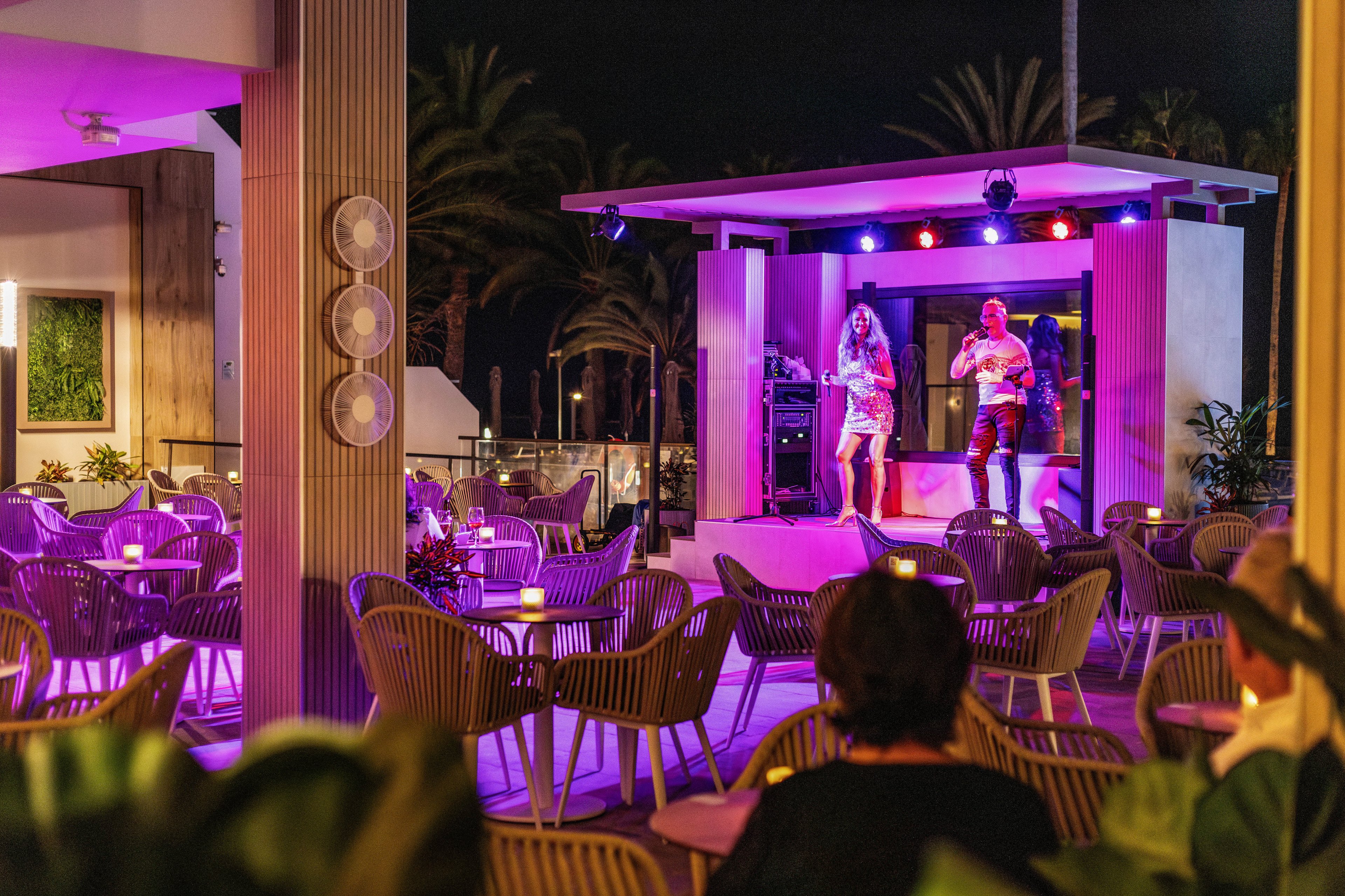 Two performers are singing on a small stage under colorful lights in an outdoor lounge setting. The area features modern chairs and tables, with a few guests seated and enjoying the ambiance. The scene is illuminated by purple and pink lighting, creating a vibrant and relaxed mood. Palm trees and architectural elements are visible in the background, enhancing the tropical atmosphere.