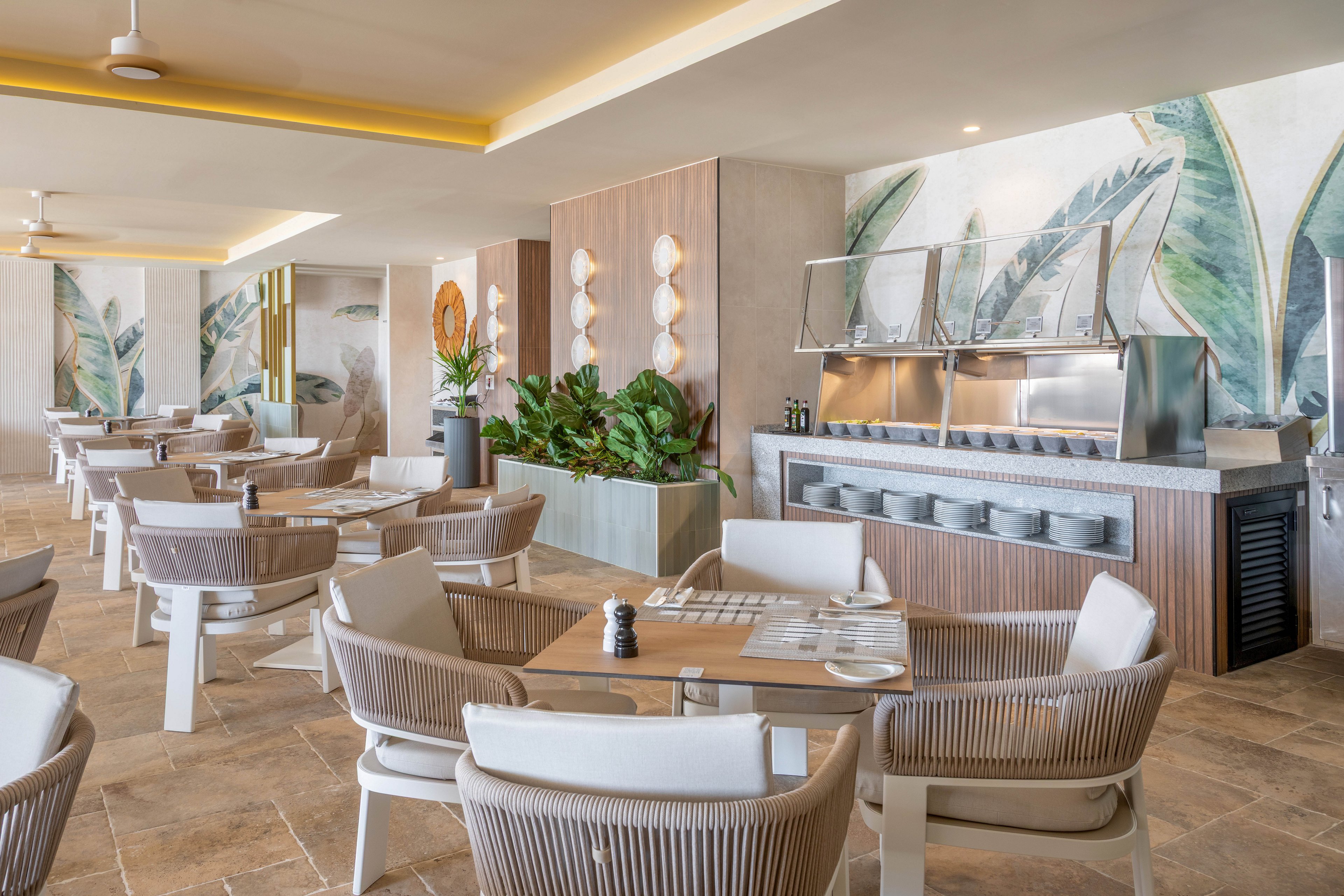 A spacious, contemporary dining area features light wood furniture and woven chairs arranged around set tables. The setting includes a buffet counter with covered serving trays and a backdrop of large tropical leaf wall art. Natural light, neutral tones, and lush green plants create a relaxed, inviting atmosphere.
