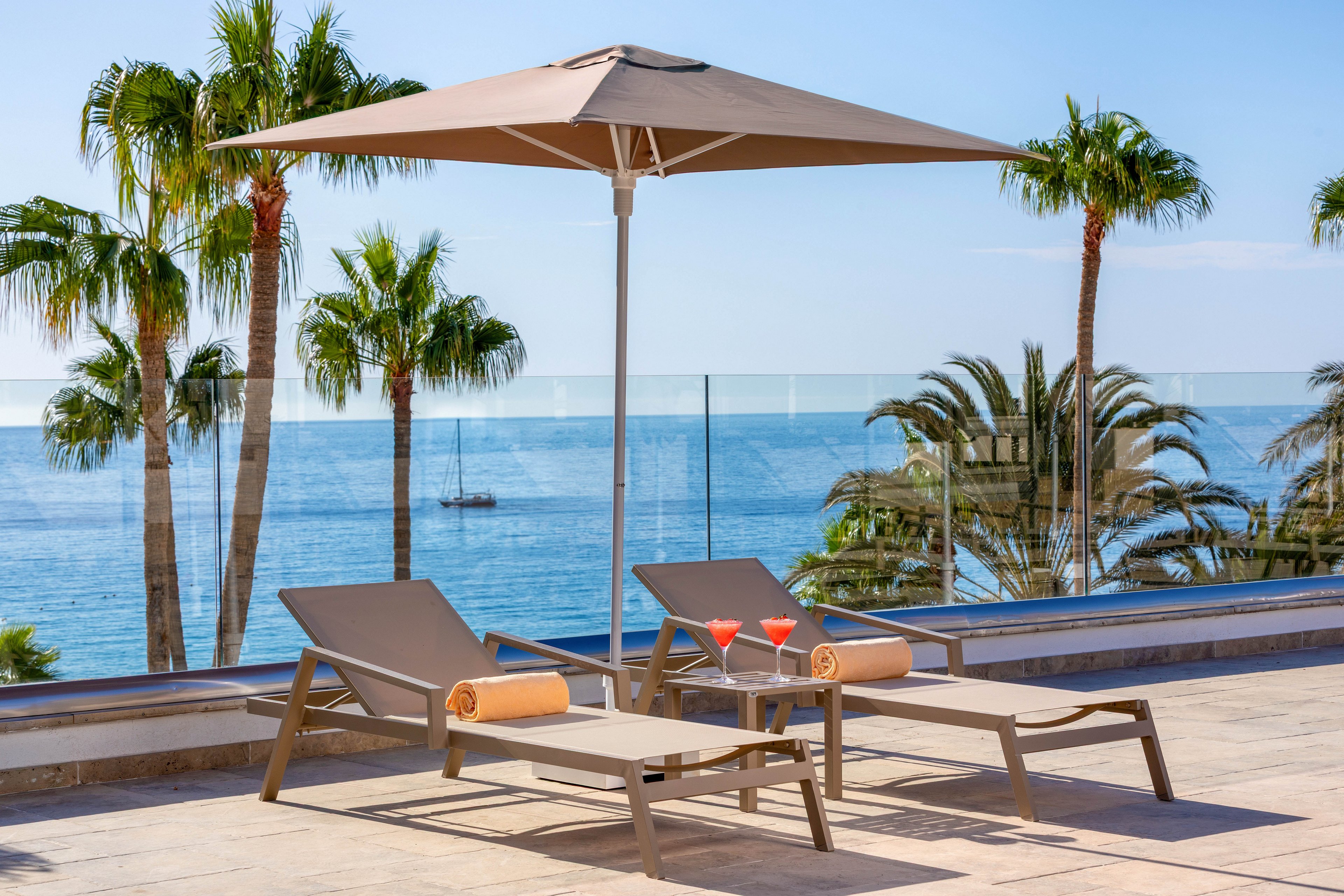 Two modern lounge chairs with rolled towels sit beneath a large umbrella on a sunlit terrace. The setting features a stunning ocean view with palm trees and a glass railing, creating a relaxing atmosphere. Two red cocktails are placed on a small table between the chairs, adding a vibrant touch to the scene. The overall mood is tranquil and inviting, perfect for leisure and relaxation.