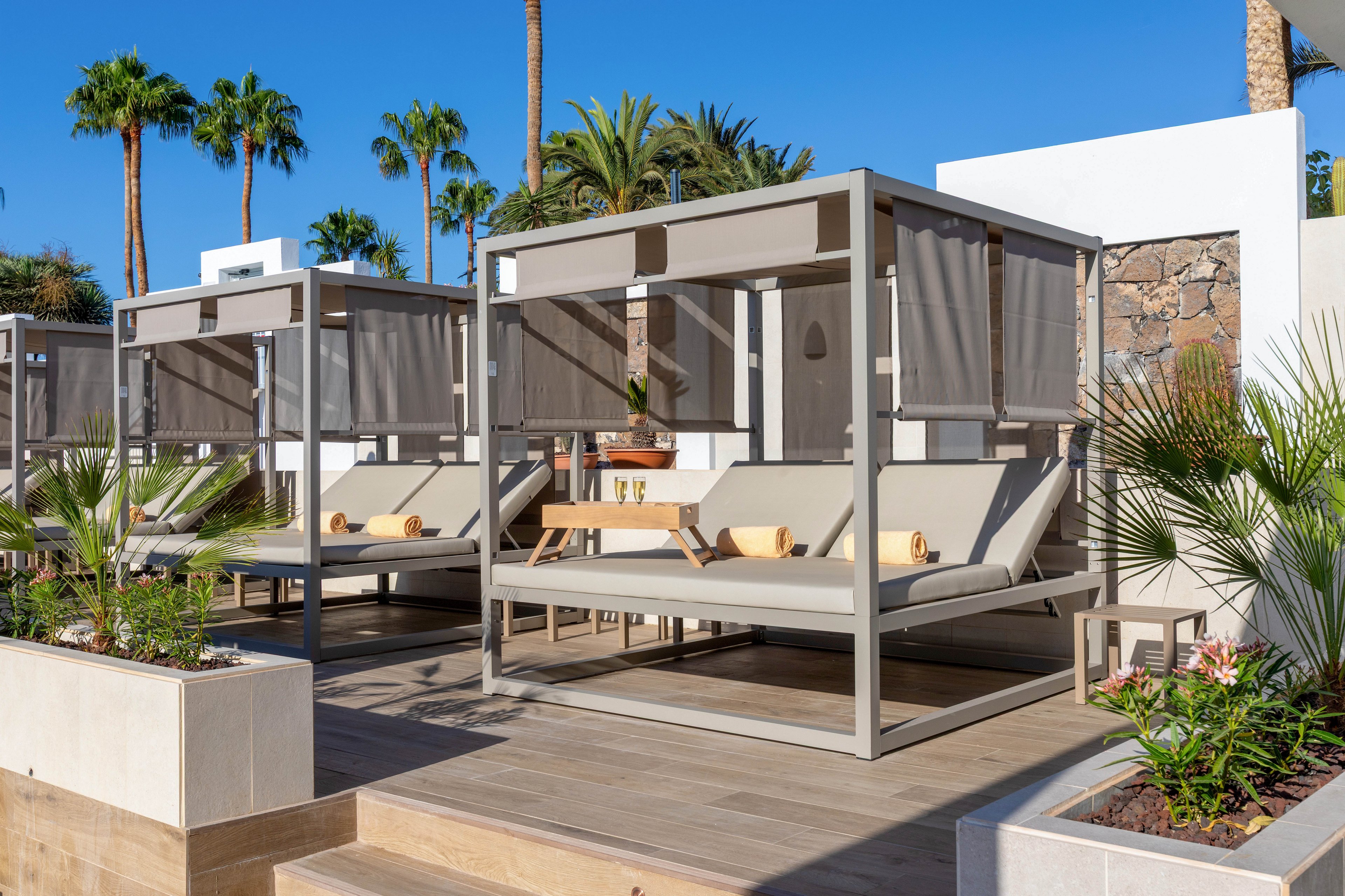 The image features stylish outdoor cabanas with cushioned sun loungers, set on a wooden deck. Rolled towels are placed on the loungers, and small tables are positioned between them. The setting is a sunny resort area with palm trees and clear blue skies, creating a relaxed and inviting atmosphere. Neutral tones and contemporary design elements dominate the scene.