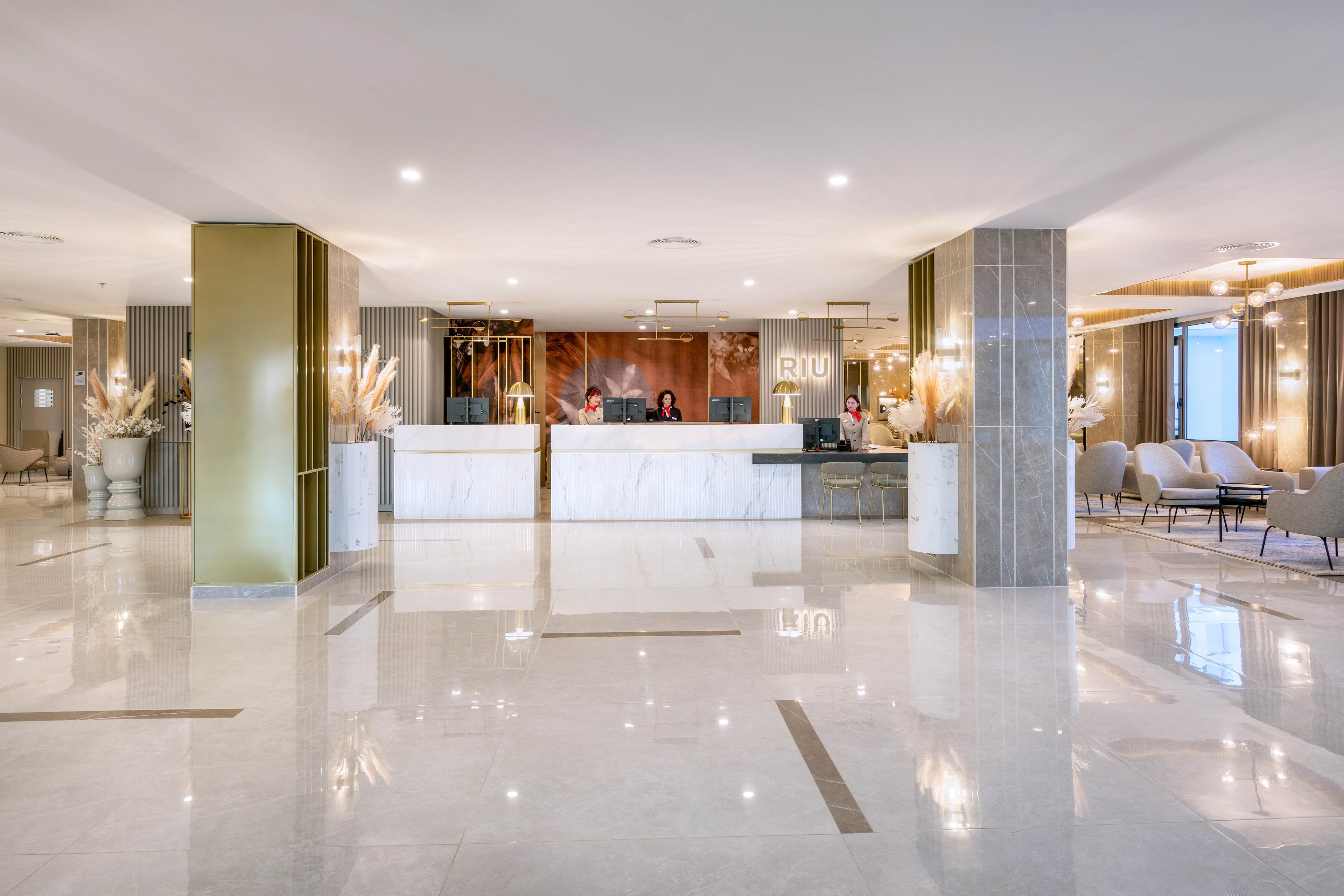 The image shows a spacious hotel lobby featuring a white marble reception desk with the RIU logo clearly visible. Two staff members are present behind the desk, welcoming guests. The setting is bright and contemporary, with neutral tones, elegant seating areas, and decorative plants. The polished floor reflects the lighting, contributing to a clean and inviting atmosphere.