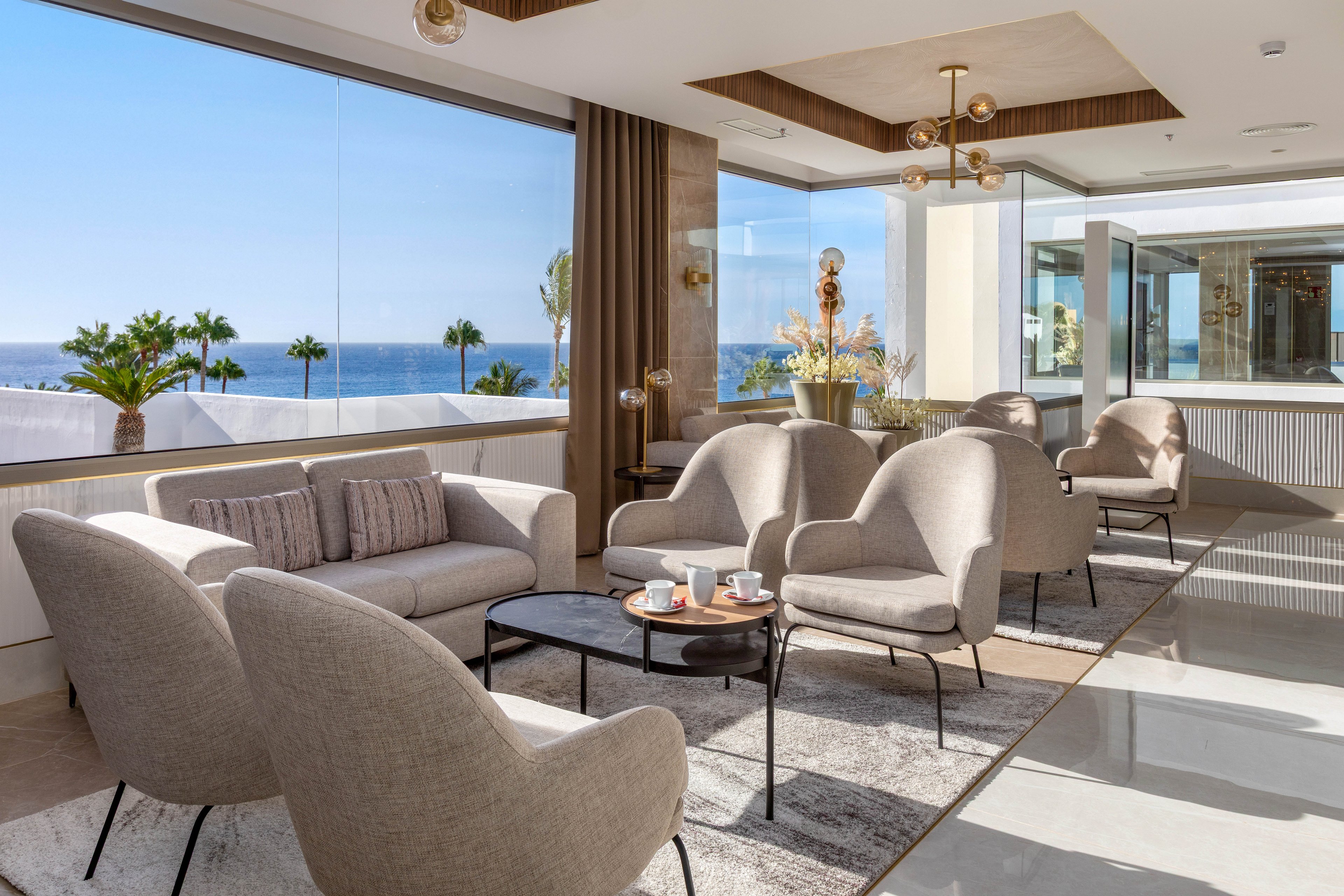 A contemporary lounge area features plush beige armchairs and a sofa arranged around black coffee tables. Large windows offer a panoramic ocean view, with palm trees visible outside. The space is bright and airy, accented by minimalist decor, neutral tones, and natural light. Several cups and plates are placed on the tables, suggesting a relaxed social setting.