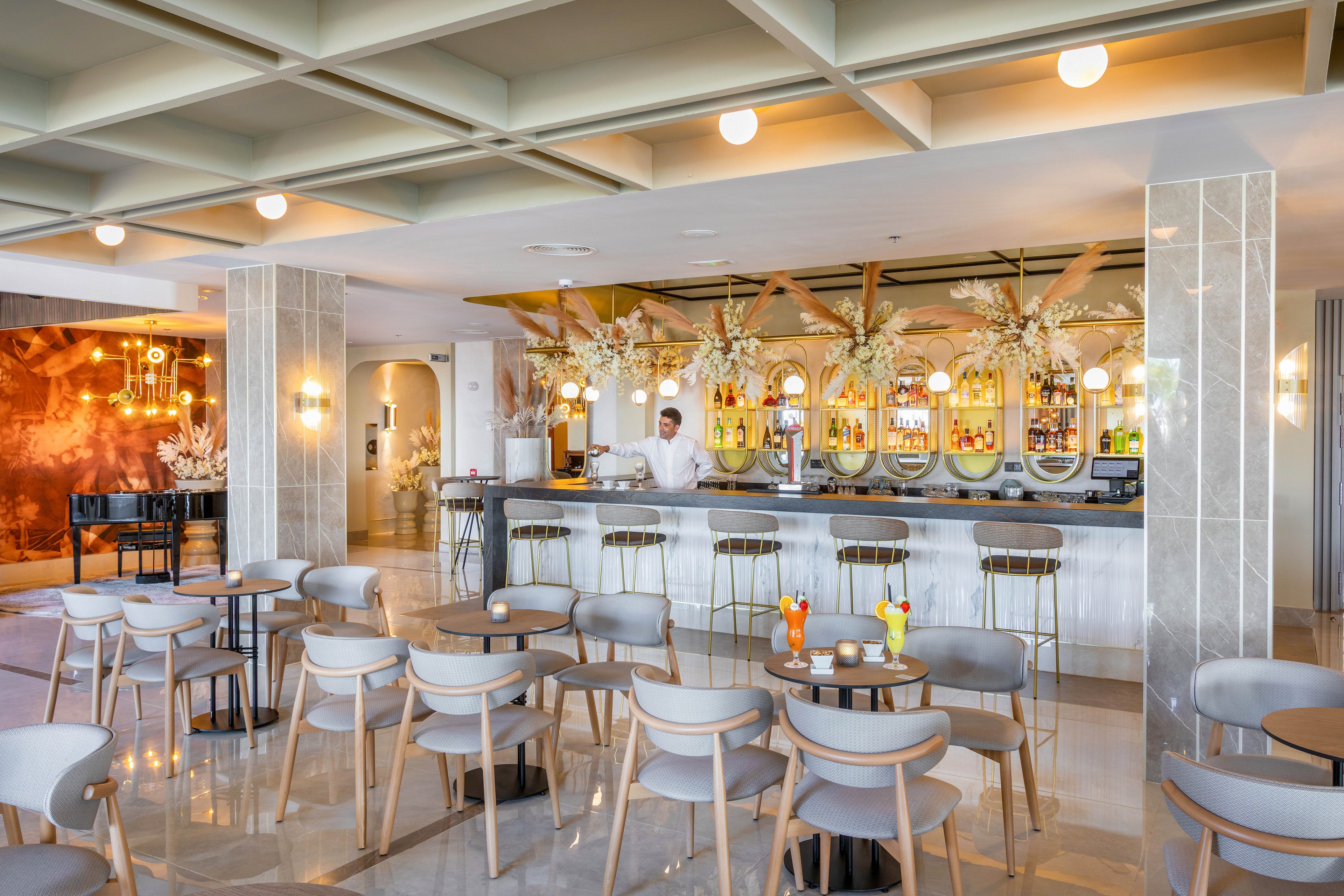 A contemporary bar area features a bartender behind a marble counter, surrounded by elegant chairs and tables. The setting is bright and airy, with neutral tones and decorative lighting. Several colorful cocktails are placed on the tables, and floral arrangements adorn the bar. The mood is sophisticated and welcoming, ideal for social gatherings or relaxation.