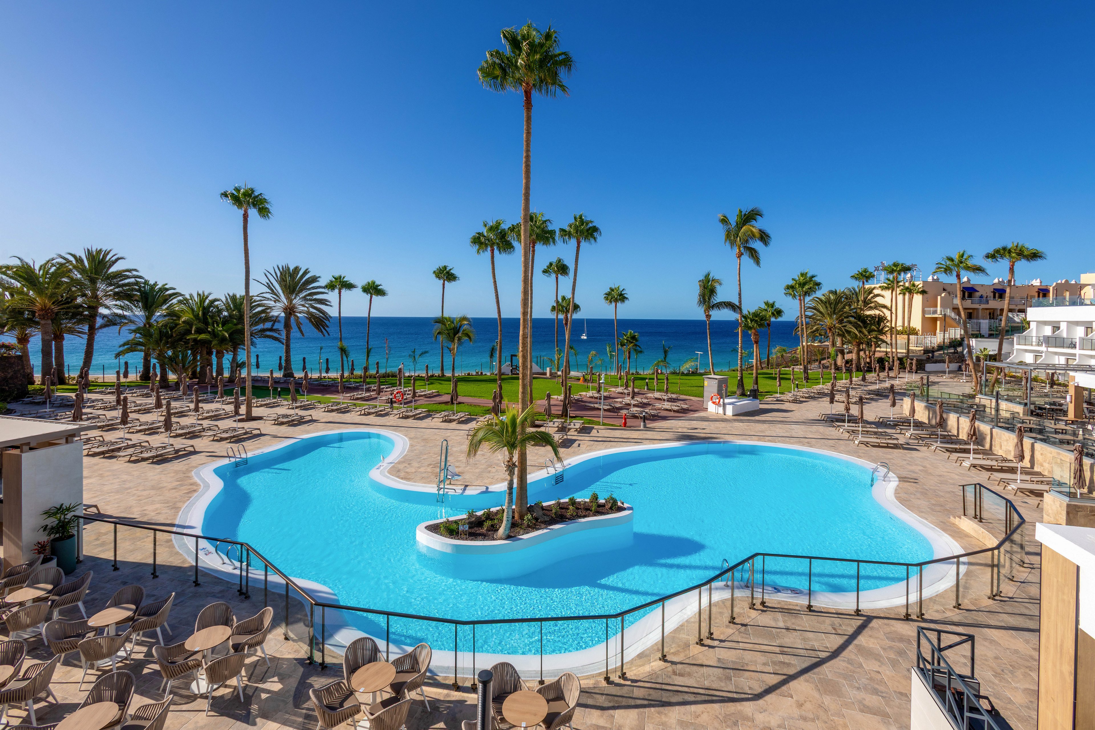 A large, uniquely shaped swimming pool is the central focus of this resort scene, surrounded by sun loungers and palm trees. The setting overlooks a clear blue ocean under a bright, cloudless sky, creating a tranquil and inviting atmosphere. The area is well-maintained, with clean poolside seating and lush greenery, suggesting a relaxing vacation destination. No visible text or numbers are present in the image.