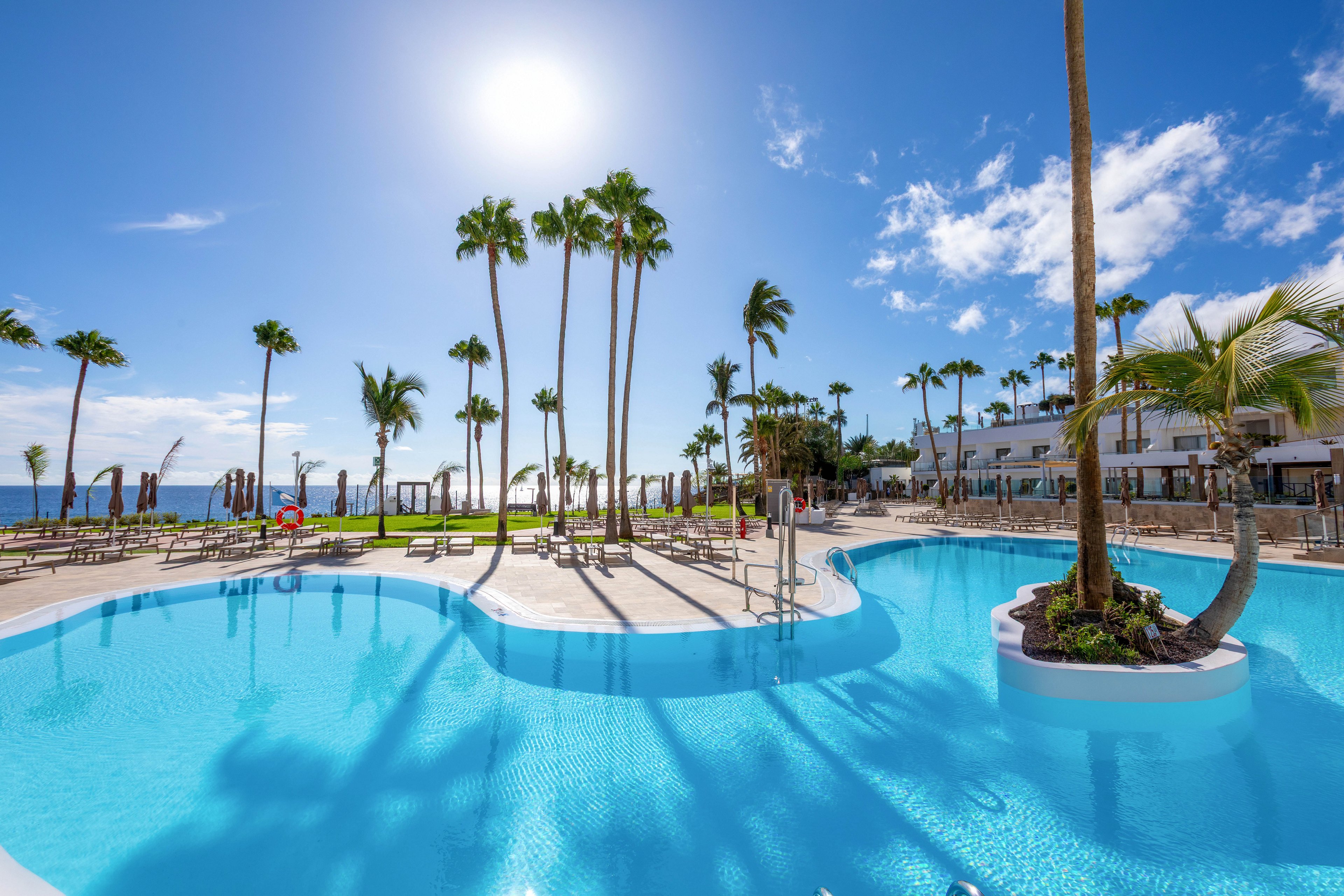 A large outdoor swimming pool is the main focus, surrounded by tall palm trees and sun loungers. The setting is a bright, sunny day with clear blue skies, and a modern white hotel building is visible in the background. The pool water is sparkling, and the overall mood is relaxing and tropical, ideal for vacation or leisure.