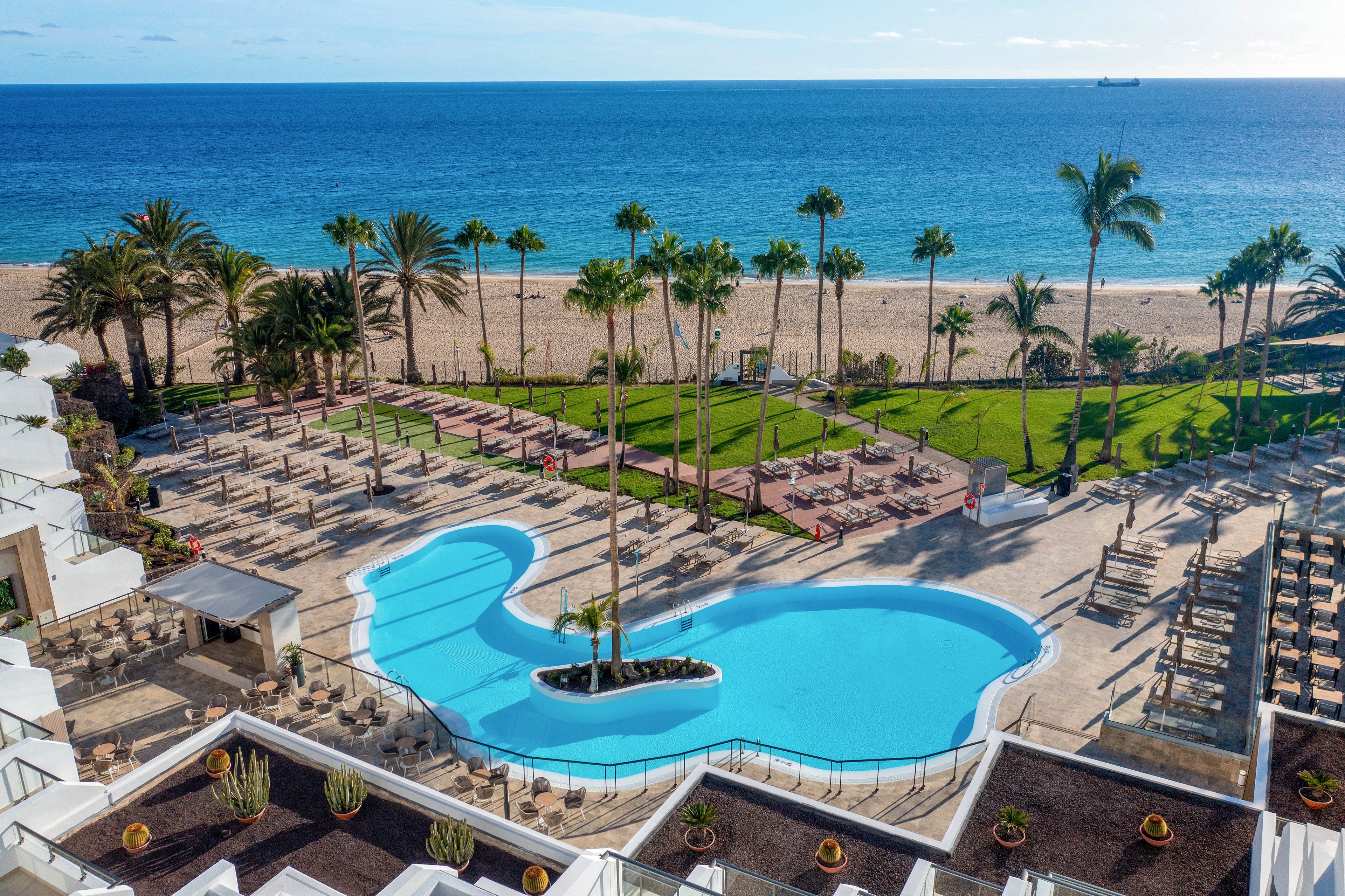 The main subject is a large, uniquely shaped swimming pool surrounded by lounge chairs and palm trees. The setting is an outdoor resort area adjacent to a sandy beach and clear blue ocean, with landscaped gardens and sunlit terraces. The visuals feature vibrant blue water, green lawns, and tropical scenery, creating a relaxed and inviting mood. No visible text or numeric values are present in the image.