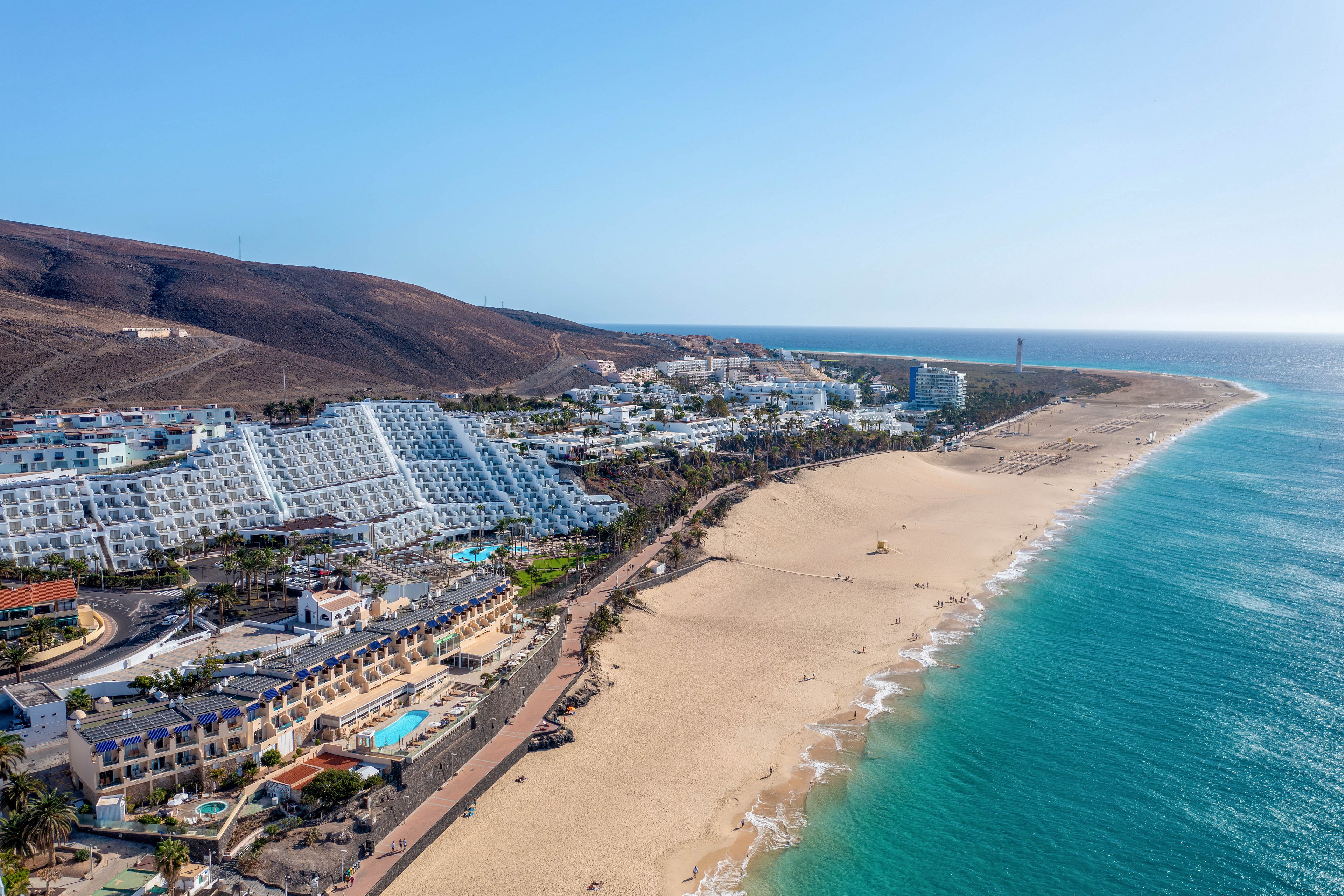 This image shows a sweeping aerial perspective of a coastal resort town with white buildings lining the shore. The scene features a wide sandy beach meeting turquoise ocean waters, with a mountainous backdrop. The area appears sunny and tranquil, with a few people visible on the beach and in the water. The overall mood is relaxed, highlighting a popular vacation destination.