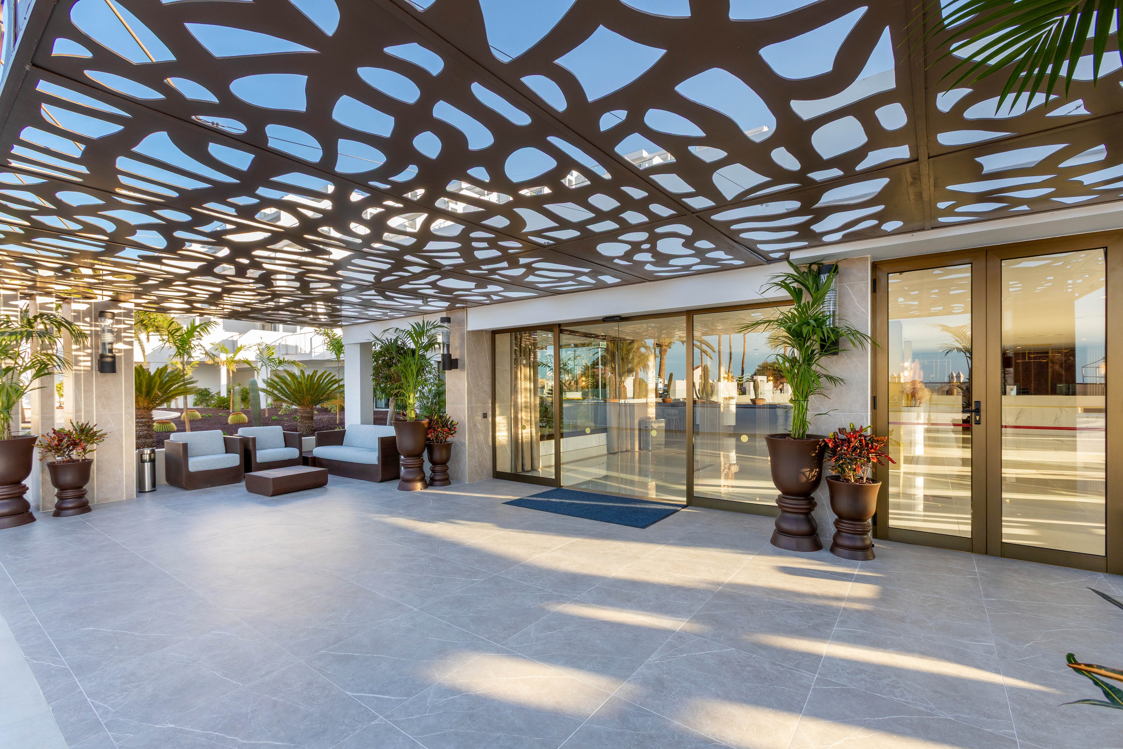 The image features a contemporary outdoor lounge area with stylish seating and large potted plants. A striking geometric-patterned canopy casts intricate shadows across the tiled floor, creating a dynamic visual effect. Glass doors lead into a bright interior space, and the overall palette is neutral with touches of greenery. No visible text or numeric values are present in the scene.