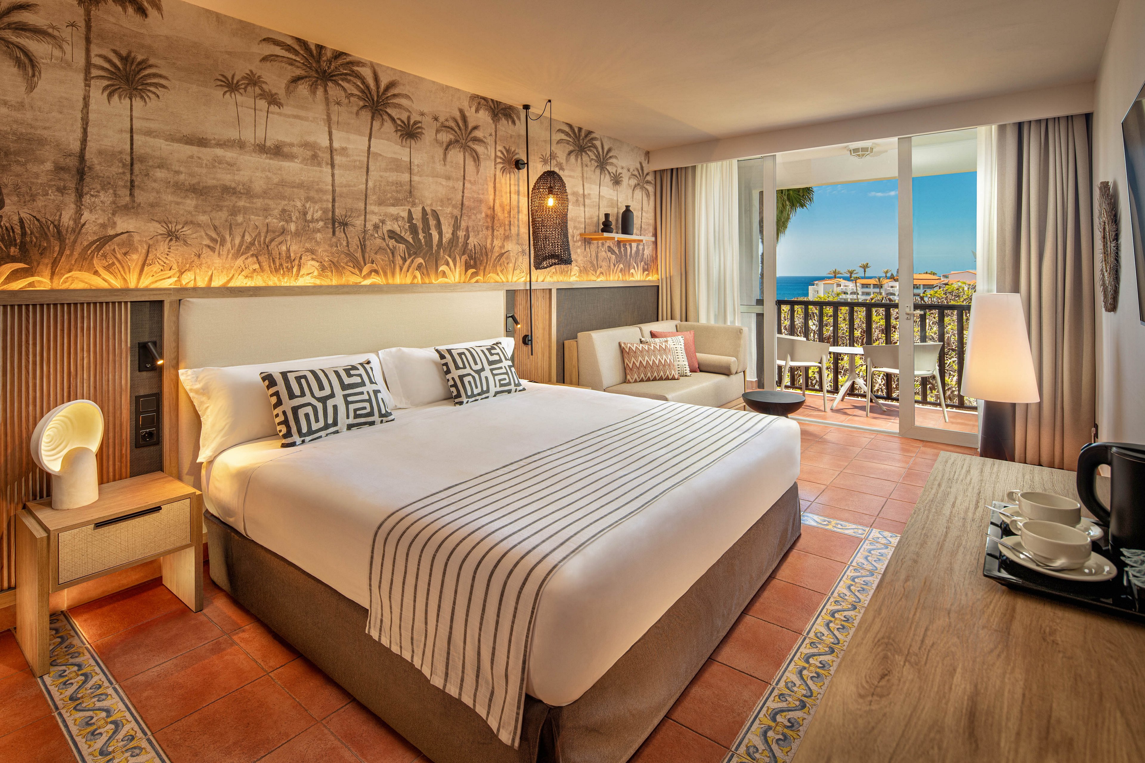 A stylish hotel bedroom features a large bed with patterned pillows and a striped throw. Warm lighting highlights a mural of palm trees above the headboard, creating a tropical ambiance. A cozy seating area and desk with a kettle set are arranged along the side of the room. Floor-to-ceiling glass doors open to a balcony overlooking palm trees and a distant ocean view, framed by curtains.