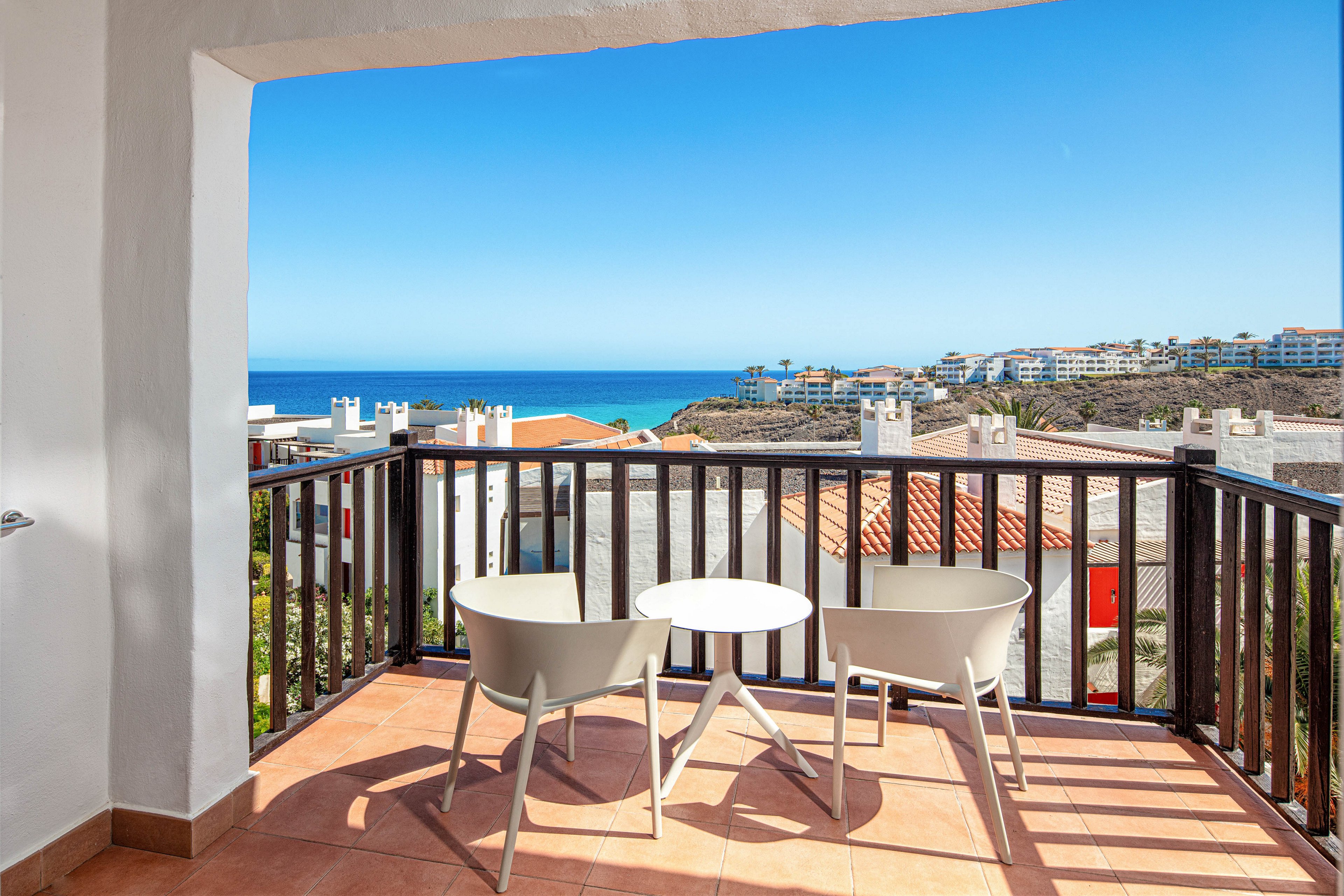 A bright balcony with two modern chairs and a small round table overlooks a coastal town. The scene features terracotta floor tiles, a dark wooden railing, and white stucco walls framing the view. In the distance, blue ocean water meets a clear sky, with low-rise buildings and rooftops visible along the shoreline. Strong sunlight casts sharp shadows, creating a warm and inviting holiday atmosphere.