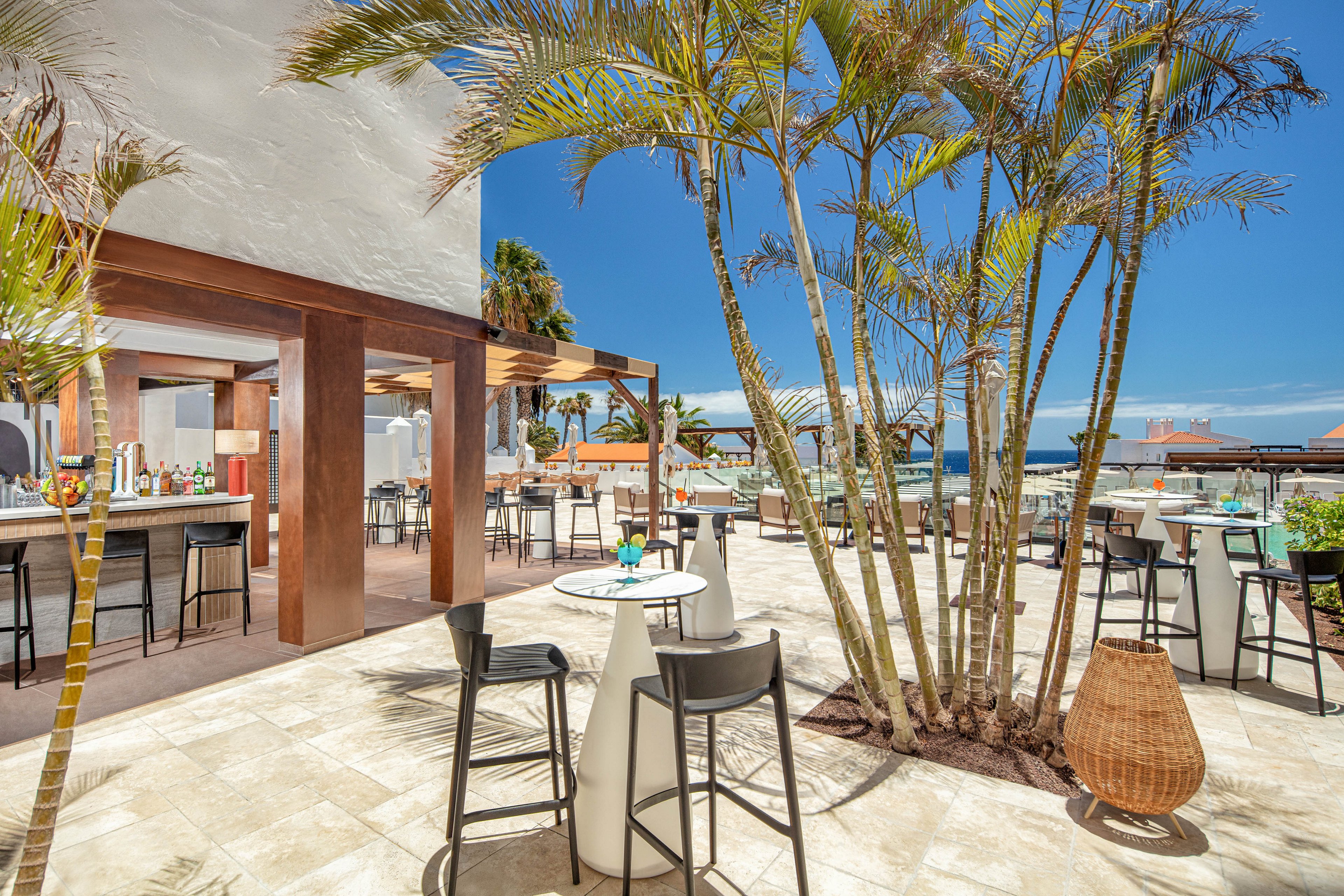 An open-air bar and lounge terrace is set under a bright blue sky near the ocean. Tall palm trees and modern high-top tables create a relaxed coastal atmosphere. Bar stools and dining chairs are arranged around small tables with drinks and decor. The scene suggests a resort-style setting ideal for casual socializing and seaside refreshments.