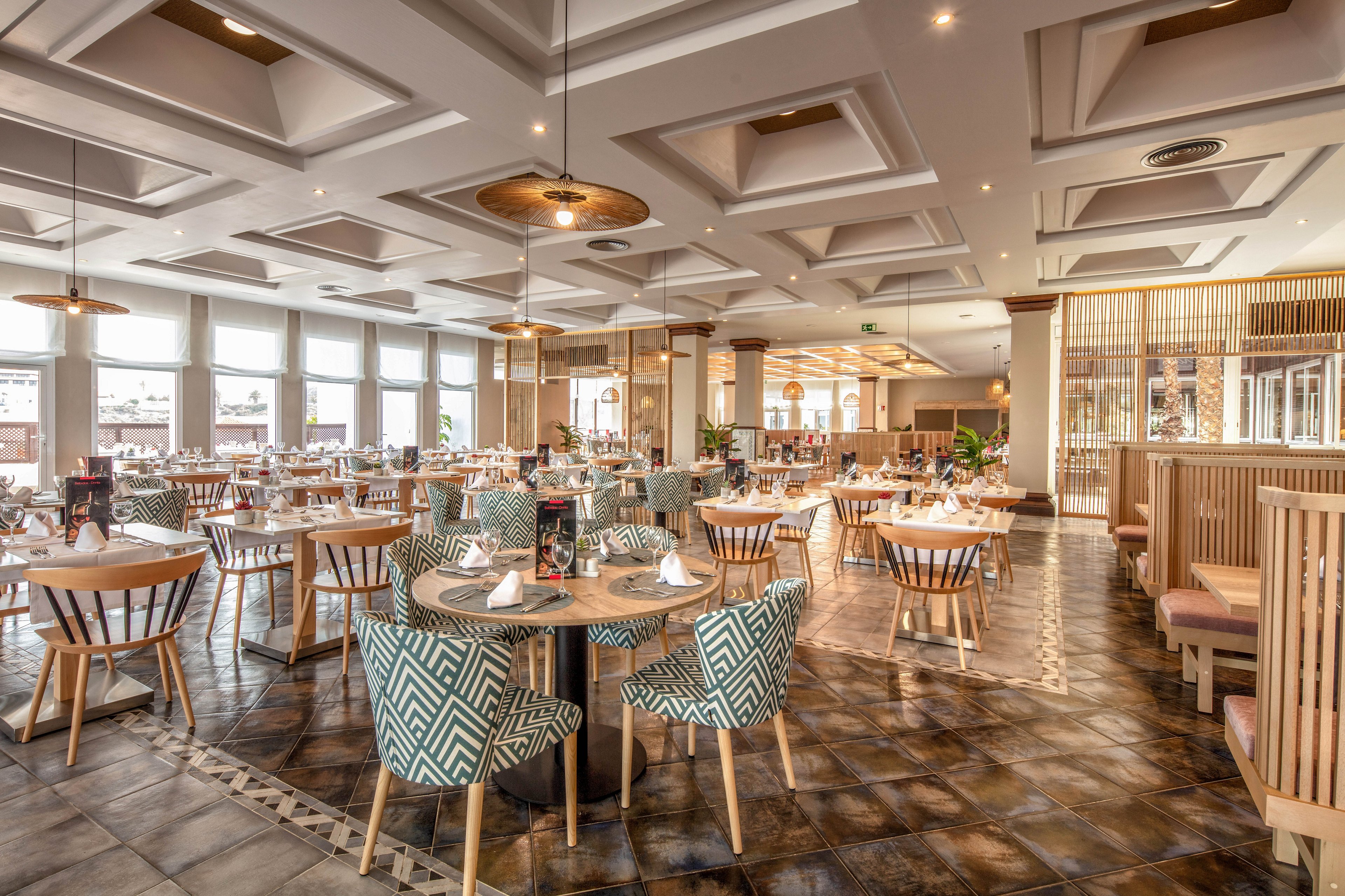 Spacious restaurant interior featuring neatly arranged round and square dining tables. The room is filled with natural light from large windows along one side, highlighting patterned upholstered chairs and wooden seating. Ceiling recess lighting and pendant lamps create a warm, inviting atmosphere. The overall design blends modern geometric details with neutral tones and clean lines for a sophisticated dining experience.