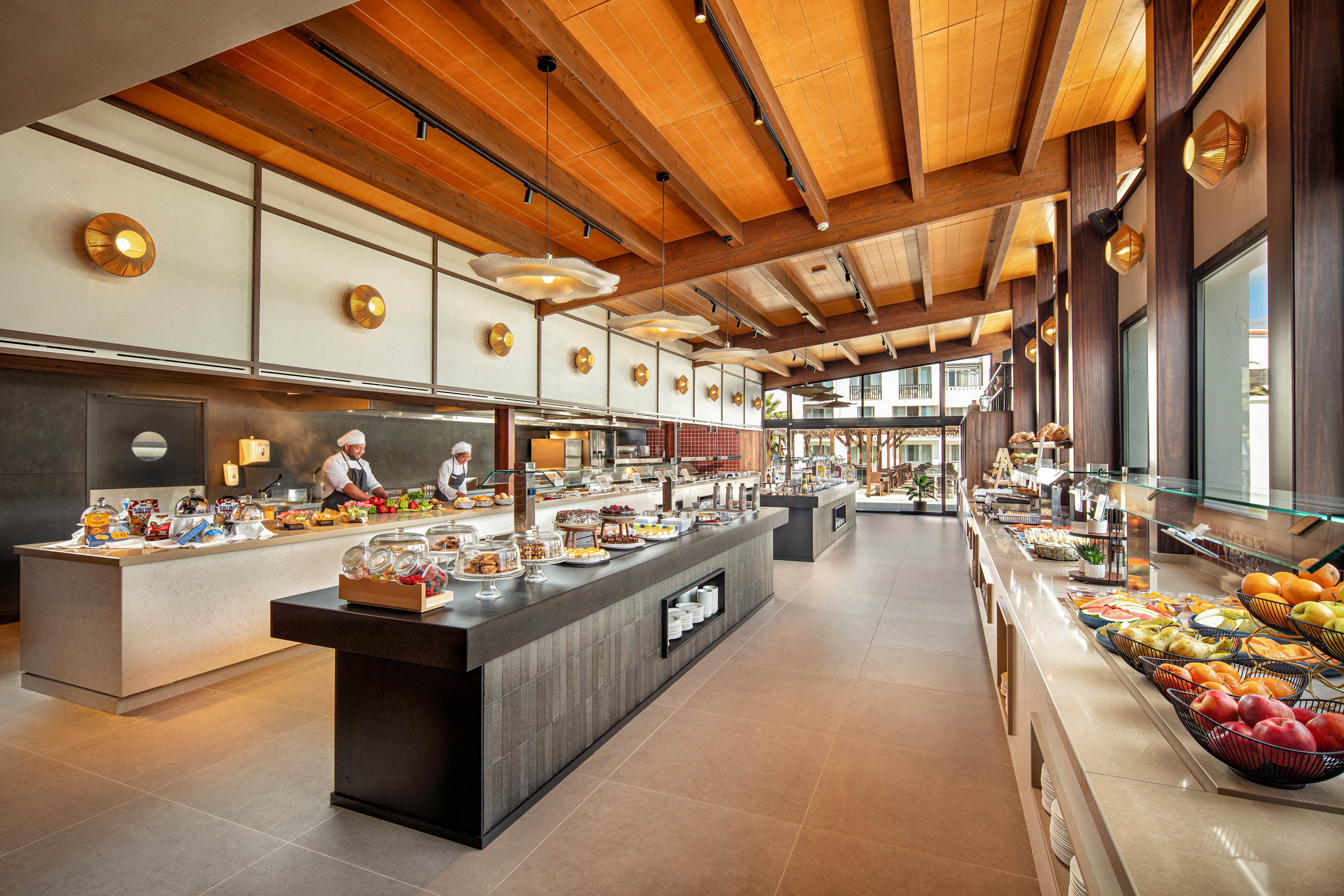A spacious, contemporary buffet restaurant features long counters filled with assorted dishes and fresh fruit. Two chefs in white uniforms and hats prepare food behind the central island. Warm wooden ceilings, large windows, and soft lighting create an inviting atmosphere. The setting appears to be part of a resort or hotel dining area with a clean, organized layout.