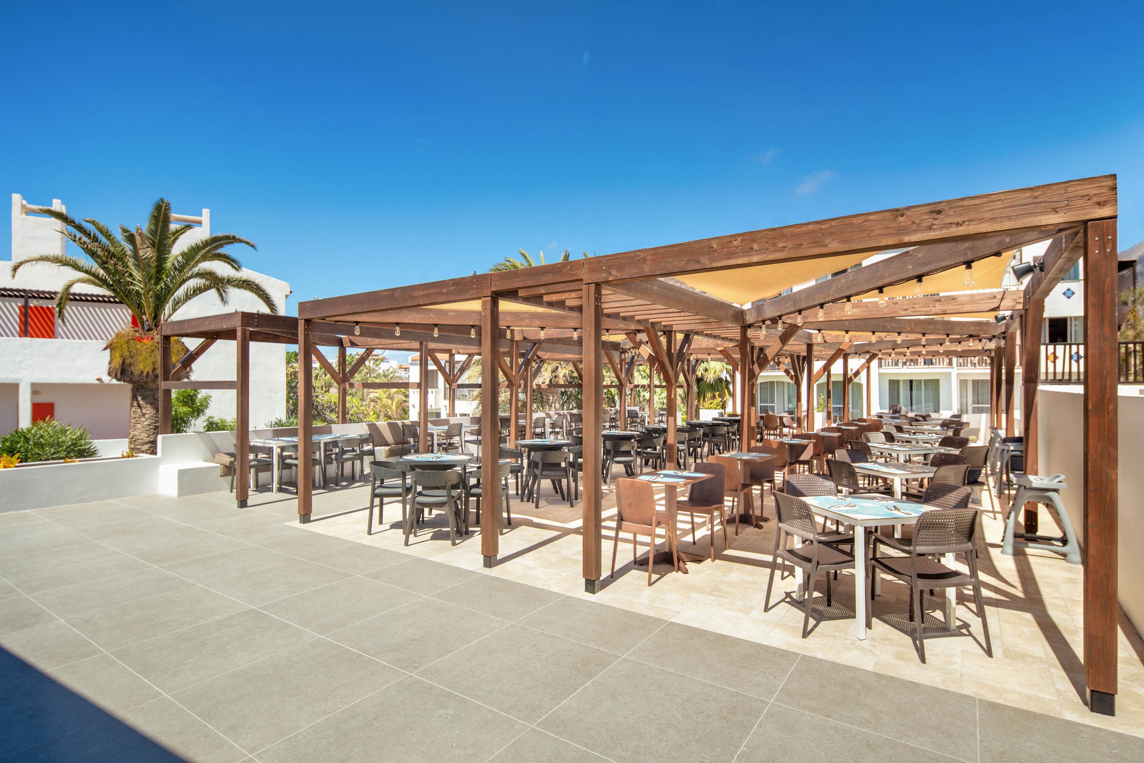 An open-air dining terrace features multiple tables and chairs arranged beneath a wooden pergola structure. The setting appears to be part of a resort or hotel complex with white buildings and balconies in the background. Bright sunlight and clear blue skies create a warm, inviting atmosphere. A palm tree and surrounding greenery add a tropical touch to the modern, tiled patio space.