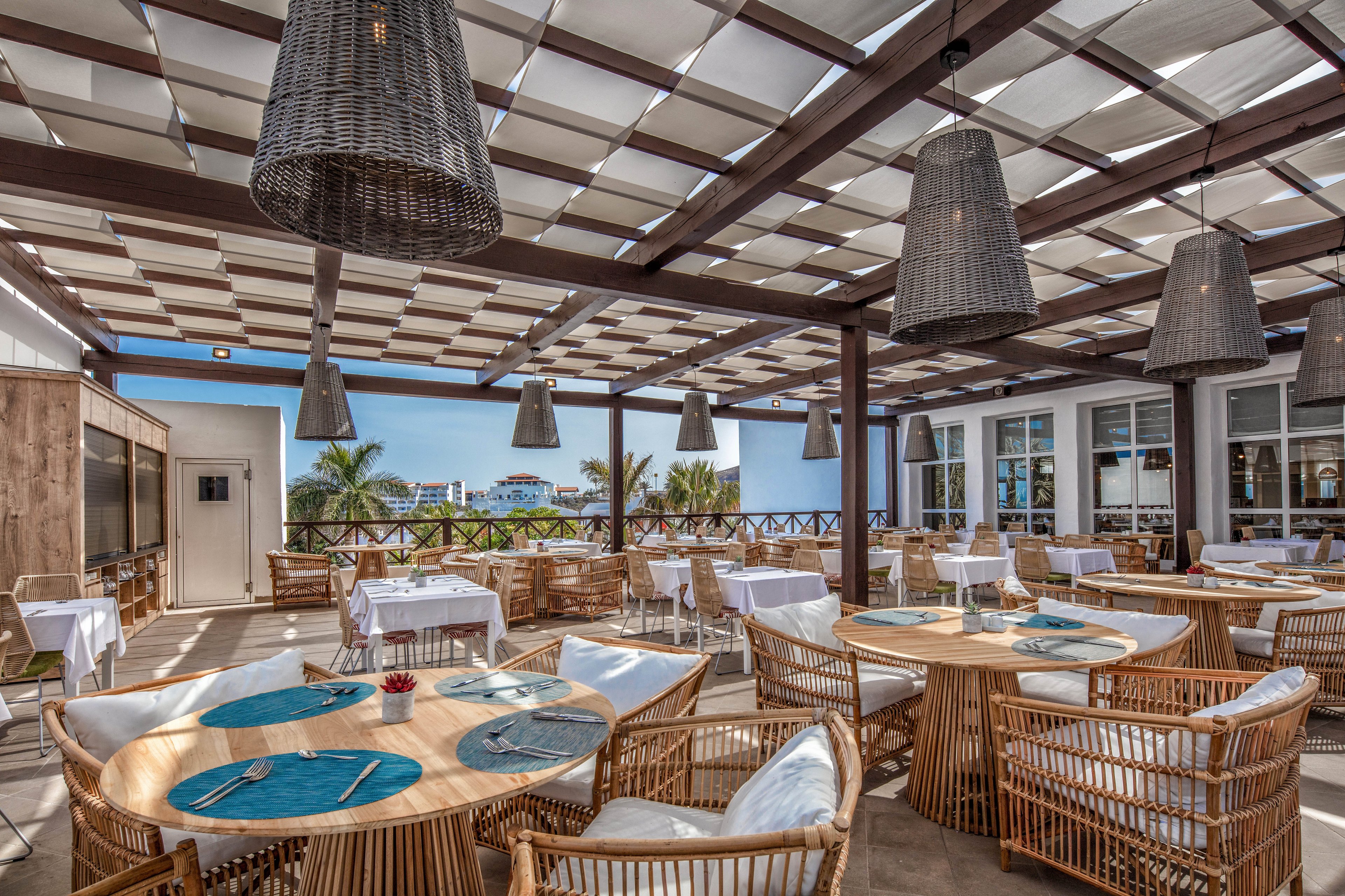 An open-air restaurant terrace features round wooden tables with wicker chairs and white cushions. Blue placemats and neatly arranged table settings create a relaxed yet refined dining atmosphere. Large woven pendant lights hang from a slatted pergola roof that filters sunlight. In the background, palm trees and distant buildings suggest a coastal or resort setting with clear blue skies.