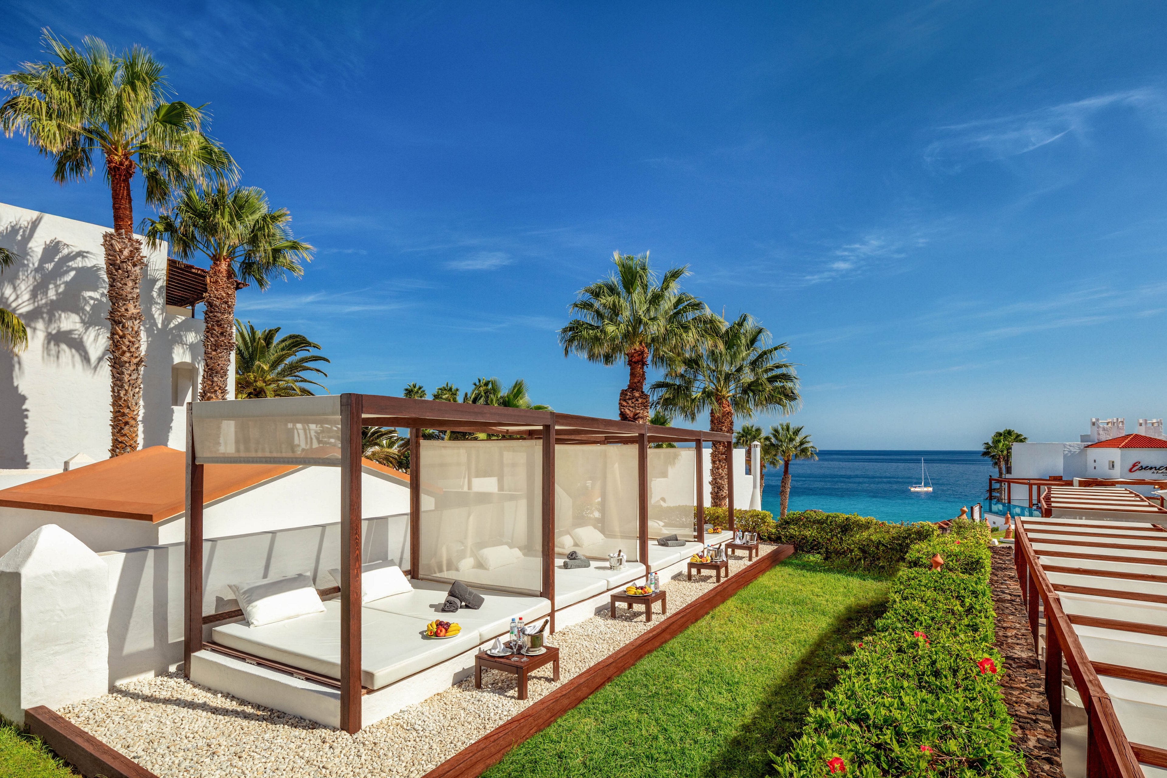 The main subject is a row of modern outdoor cabanas with white cushions and wooden frames, set in a lush garden area. The setting features palm trees, manicured lawns, and a clear blue sky, with the ocean and a sailboat visible in the background. The mood is relaxed and upscale, with a bright, sunny palette and clean architectural lines. No visible text or numeric values are present in the image.