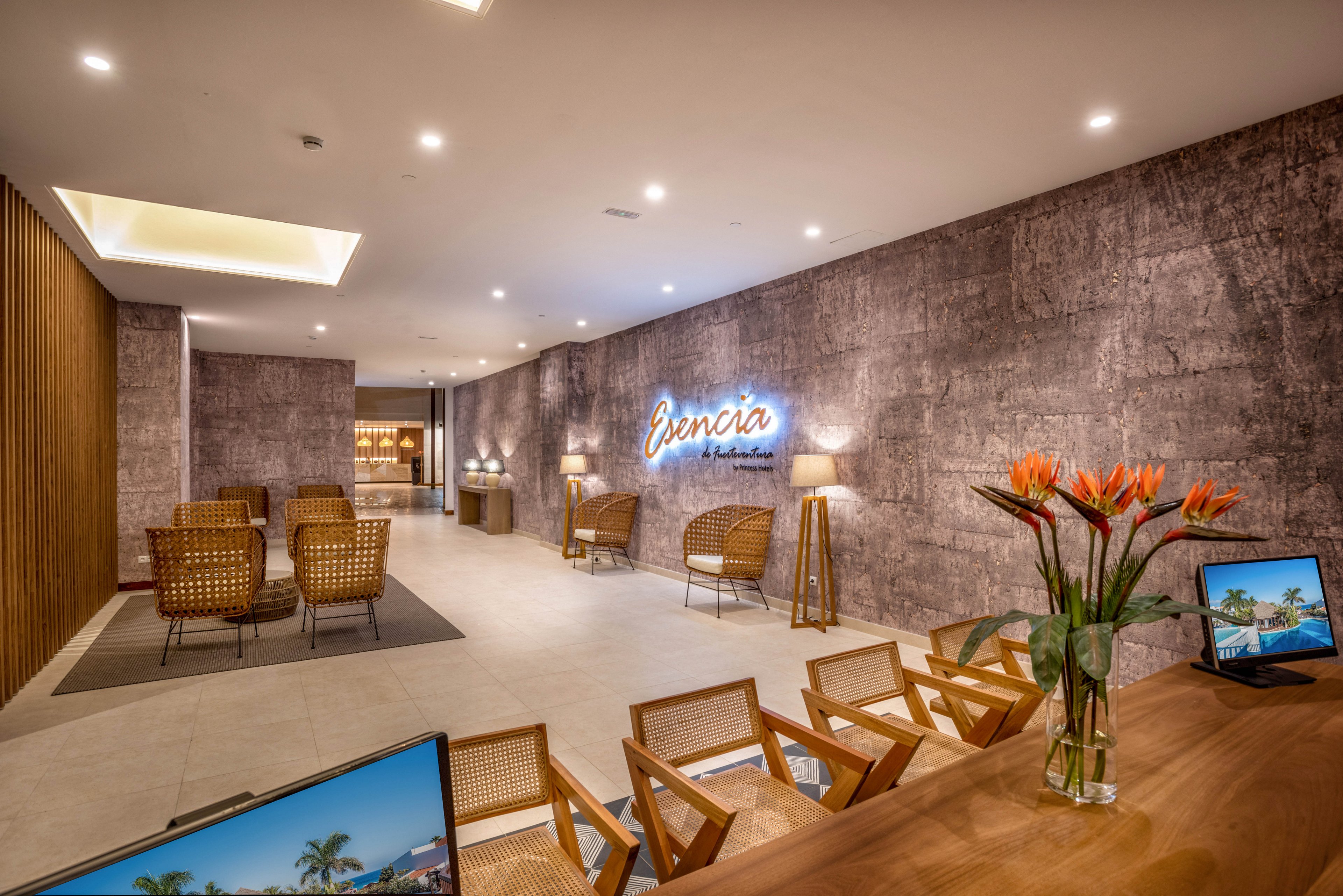 A contemporary hotel lobby features wicker chairs and wooden tables arranged for guest seating. The setting is warmly lit with a neutral palette and textured walls, highlighted by a neon sign reading 'Bencia'. Tropical flowers and computer screens displaying beach scenes add visual interest. The space exudes a welcoming and relaxed mood, suitable for leisure or business travelers.