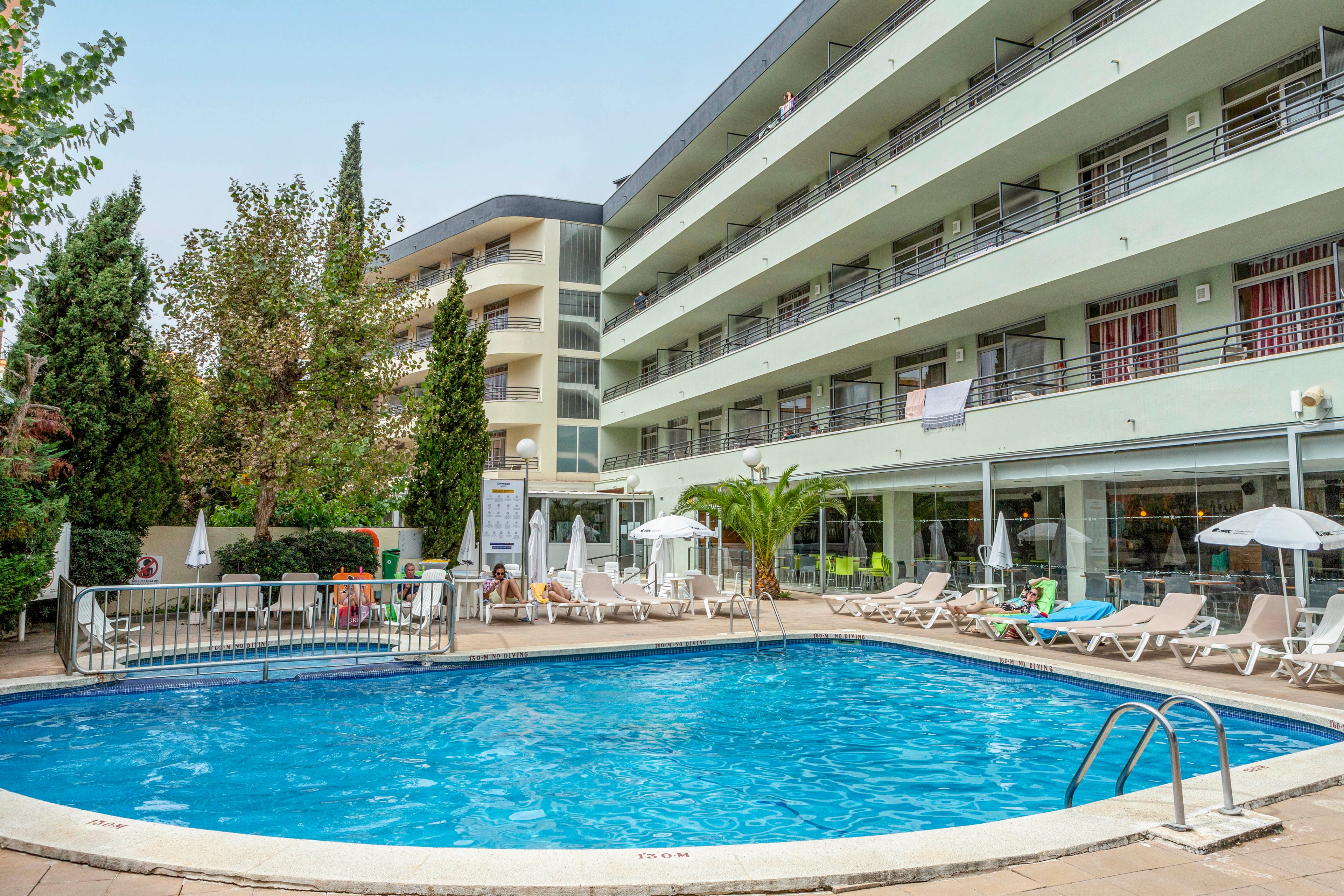 The main subject is a clear blue swimming pool surrounded by white lounge chairs and umbrellas, set in front of a modern multi-story hotel building. The scene is outdoors, featuring greenery and tall trees, with a relaxed and inviting atmosphere. The pool area is clean and well-maintained, with a few towels visible on the railings and chairs. No visible numeric values or unmistakable brand names are present.
