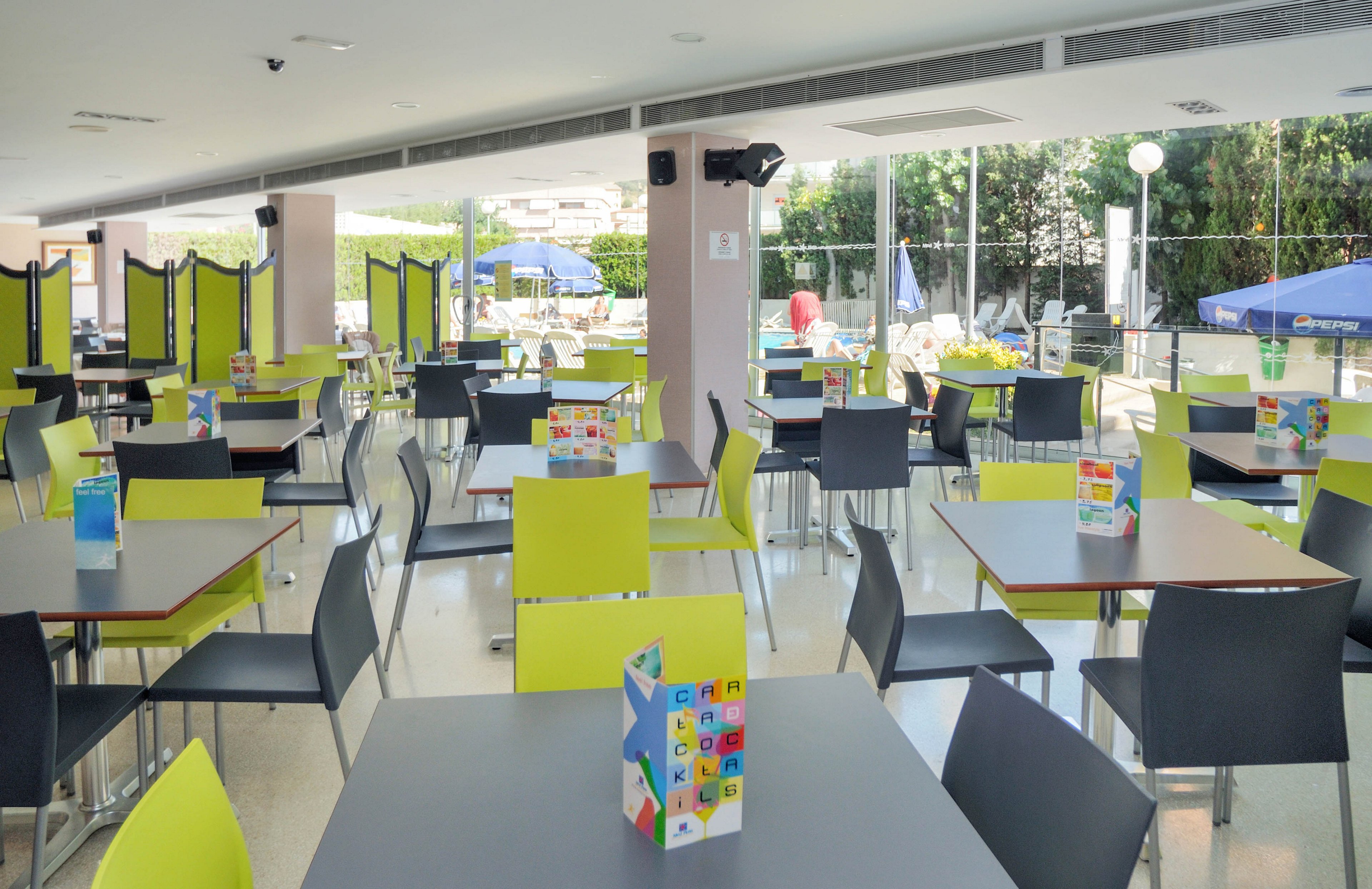 The image shows a spacious cafeteria featuring lime green and black chairs paired with dark tables. Each table has a colorful menu or information card placed upright. The setting is bright, with large windows revealing an outdoor area with umbrellas and greenery. The overall mood is clean, contemporary, and inviting, suitable for casual dining or social gatherings.