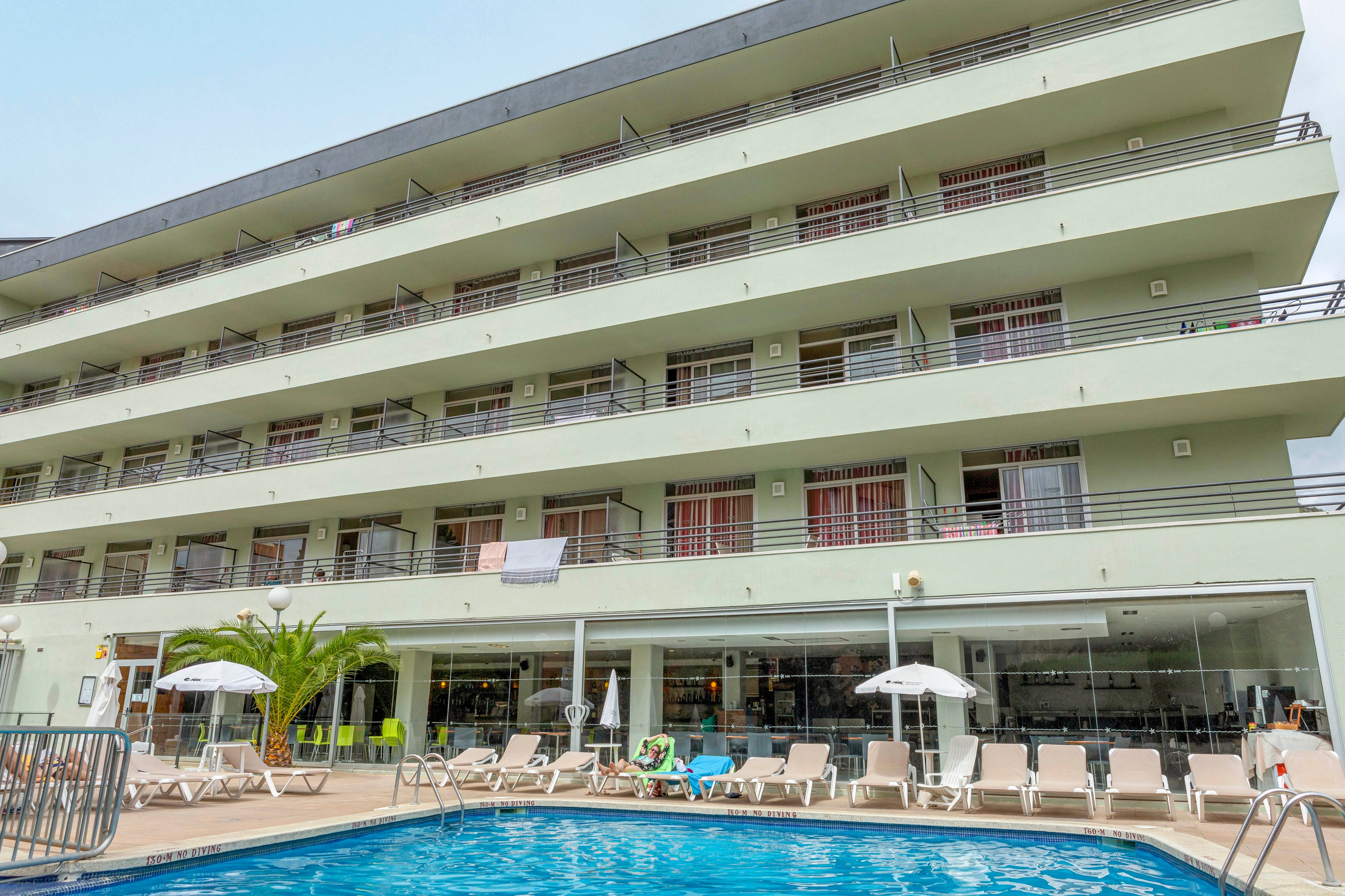 The image shows a multi-story hotel building with balconies and a ground-level pool area. Lounge chairs and umbrellas are arranged around the pool, creating a relaxing atmosphere. The setting appears to be outdoors, with palm trees and clear glass windows visible. No specific brand or numeric values are clearly identifiable in the image.