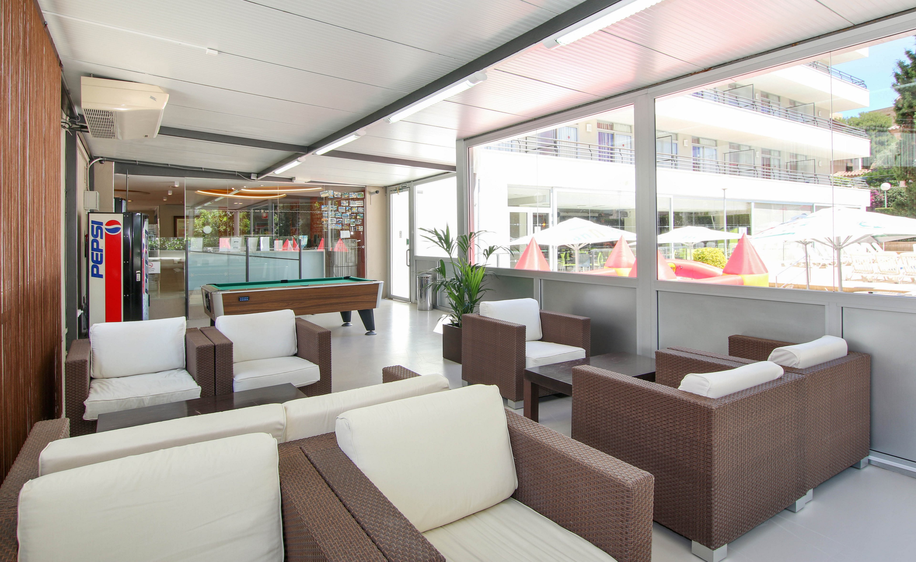 The image shows a contemporary indoor lounge featuring brown wicker armchairs with white cushions. A Pepsi vending machine is clearly visible near a pool table in the background. Large windows provide views of an outdoor area with umbrellas and a multi-story building. The setting is bright and airy, with neutral tones and clean lines.