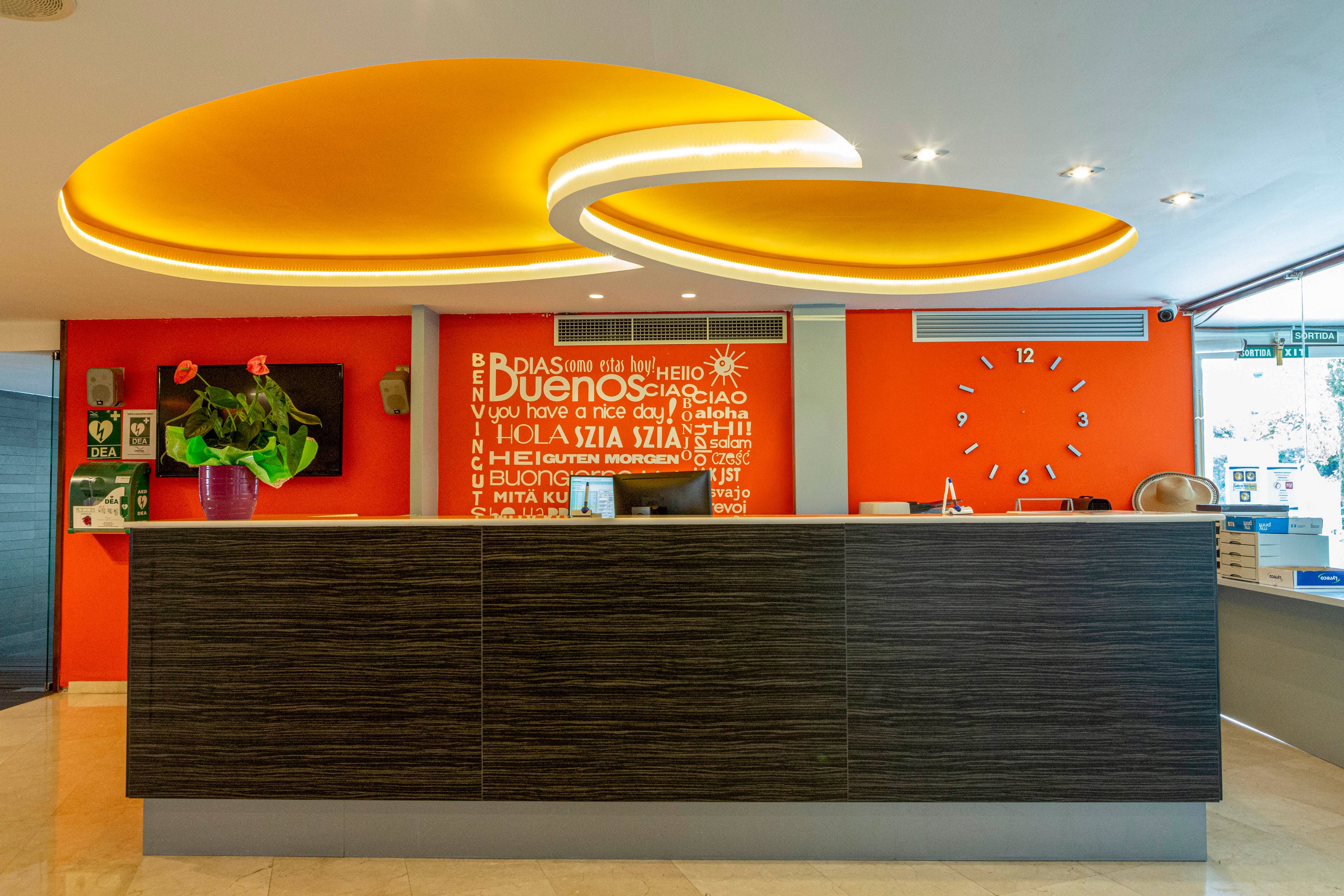 A contemporary hotel reception area features a dark wood counter and a vibrant orange accent wall. A young woman stands behind the desk, smiling, with a large orange wall clock displaying the time. The ceiling has unique, glowing yellow-orange circular light fixtures, and welcoming text in multiple languages is visible on the wall. The setting is bright and inviting, with a modern, friendly atmosphere.