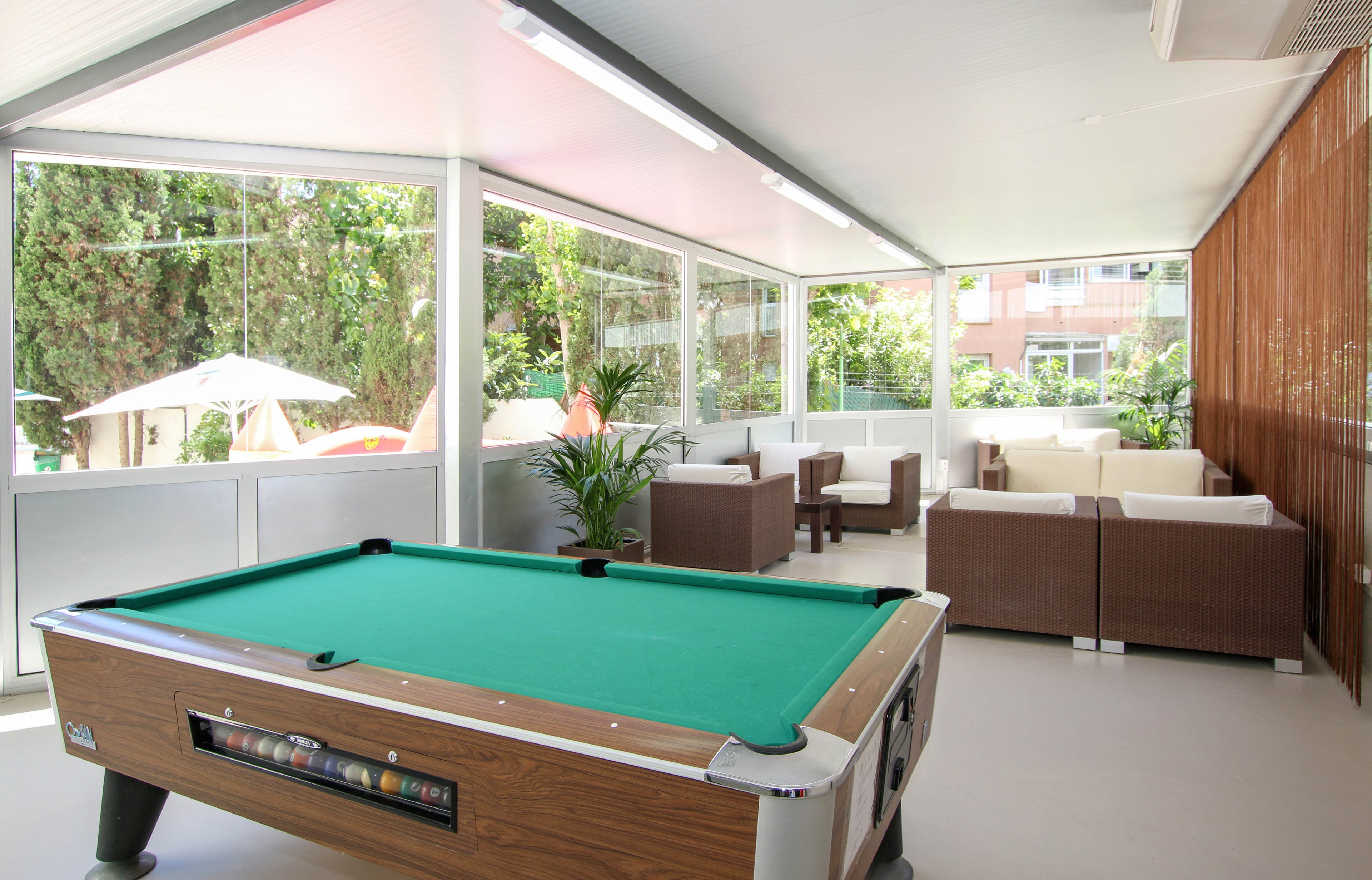 A contemporary indoor lounge features a green felt pool table as its centerpiece, surrounded by comfortable brown and white sofas. Large glass windows provide views of lush greenery and outdoor umbrellas, creating a bright and airy atmosphere. The space is well-lit with overhead lights and includes potted plants for added freshness. No visible text or numeric values are present in the image.