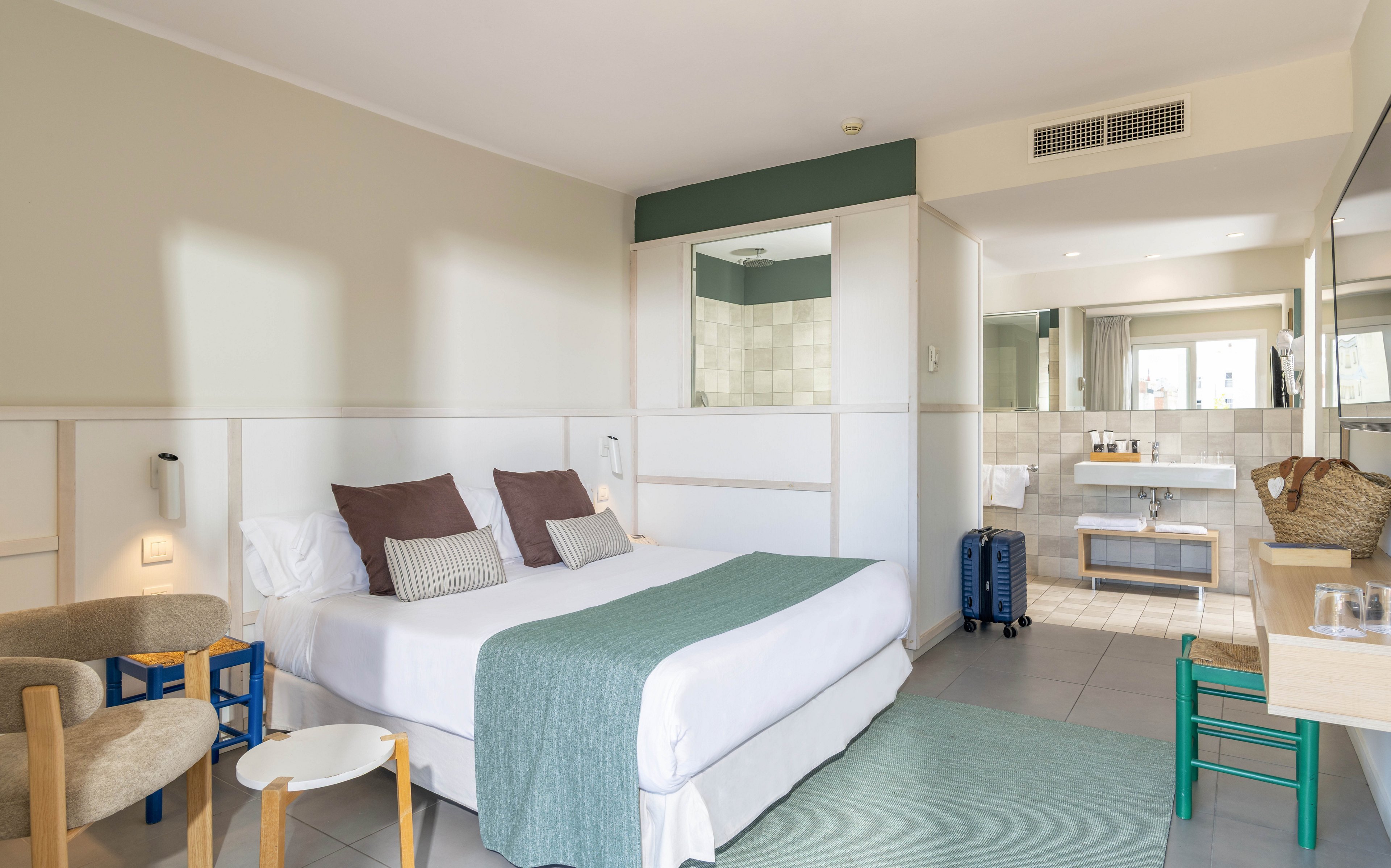 A contemporary hotel room features a neatly made double bed with white linens and a light teal throw. The space includes a small seating area with chairs and side tables, and a desk with a woven bag placed on top. An open-plan ensuite bathroom with a large mirror and vanity is visible in the background, along with a blue suitcase near the entrance. Soft neutral tones and natural light create a calm, inviting atmosphere suitable for vacation or business travel.