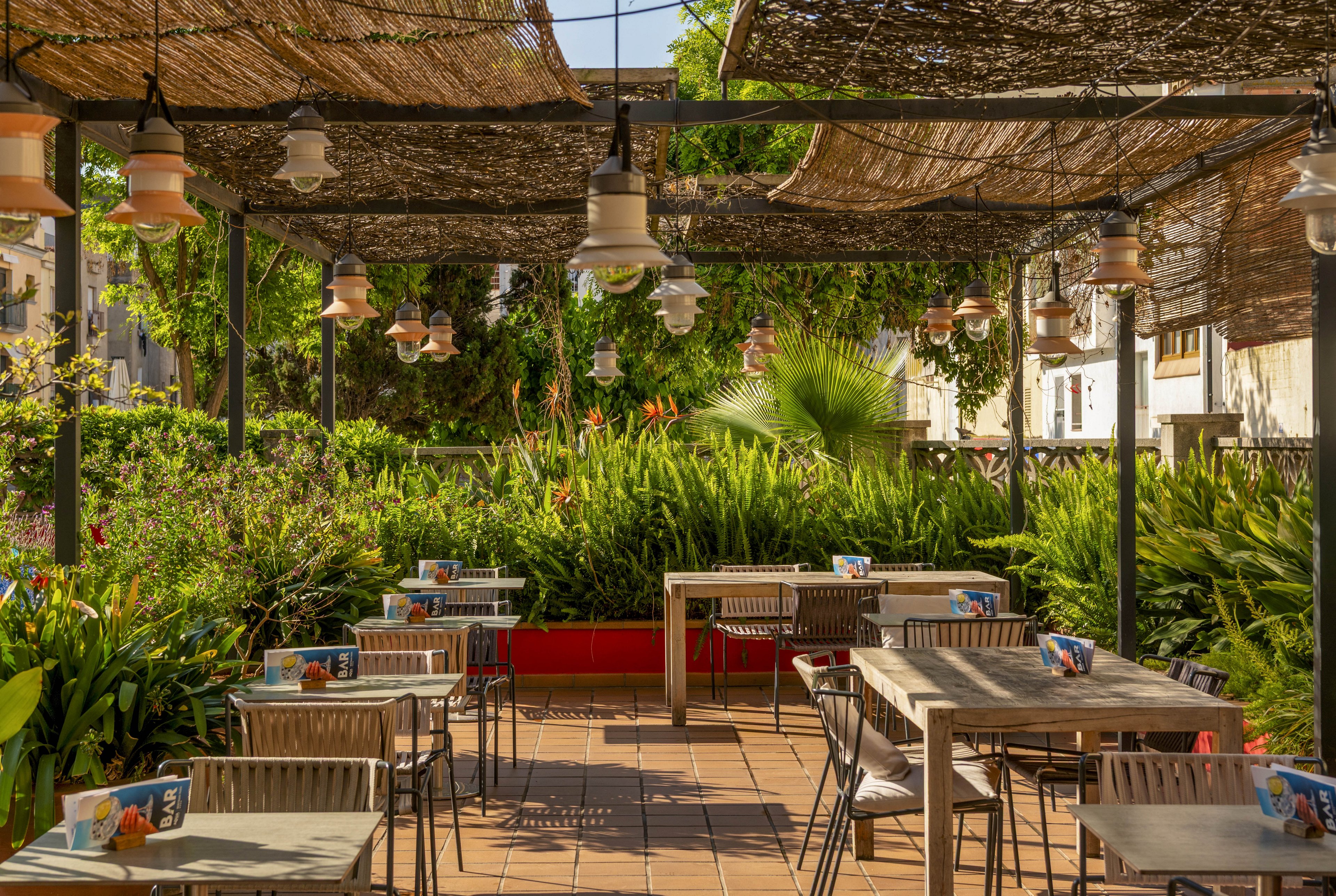 An inviting outdoor café terrace features neatly arranged tables and chairs under a woven shade canopy. The setting is surrounded by lush green plants and trees, creating a cozy garden atmosphere. Warm sunlight filters through the overhead structure, casting soft shadows on the tiled floor. Small table tents or menus sit on each table, suggesting a relaxed dining or coffee experience.