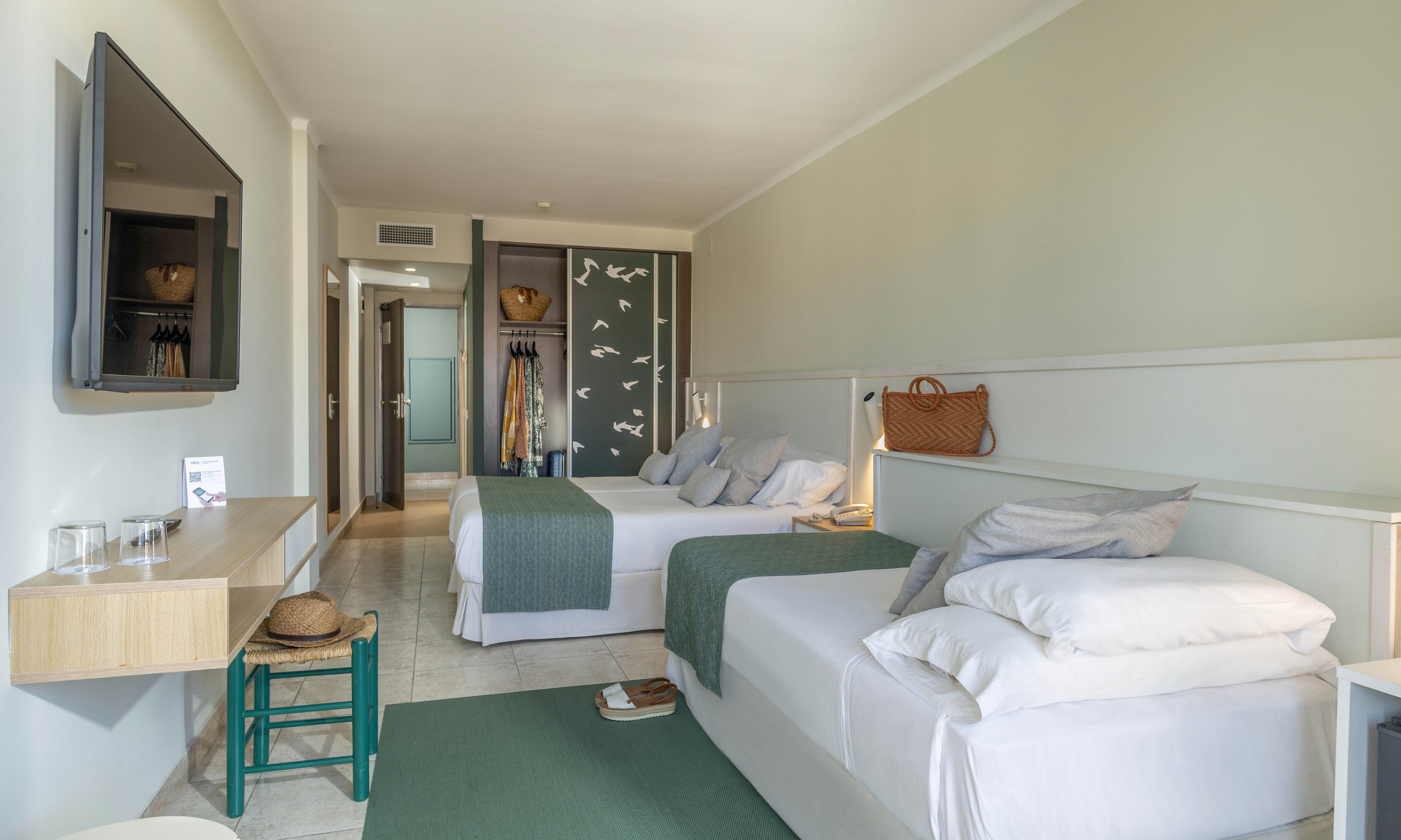A bright, modern hotel room features two neatly made beds with green throws and white linens. The space includes a wall-mounted flat-screen TV, a minimalist wooden desk, and a small green stool with a straw hat. Soft neutral walls and light flooring create an airy, coastal feel, while a decorative panel with bird silhouettes adds visual interest. A woven bag on the bed and warm bedside lighting suggest a relaxed vacation atmosphere.