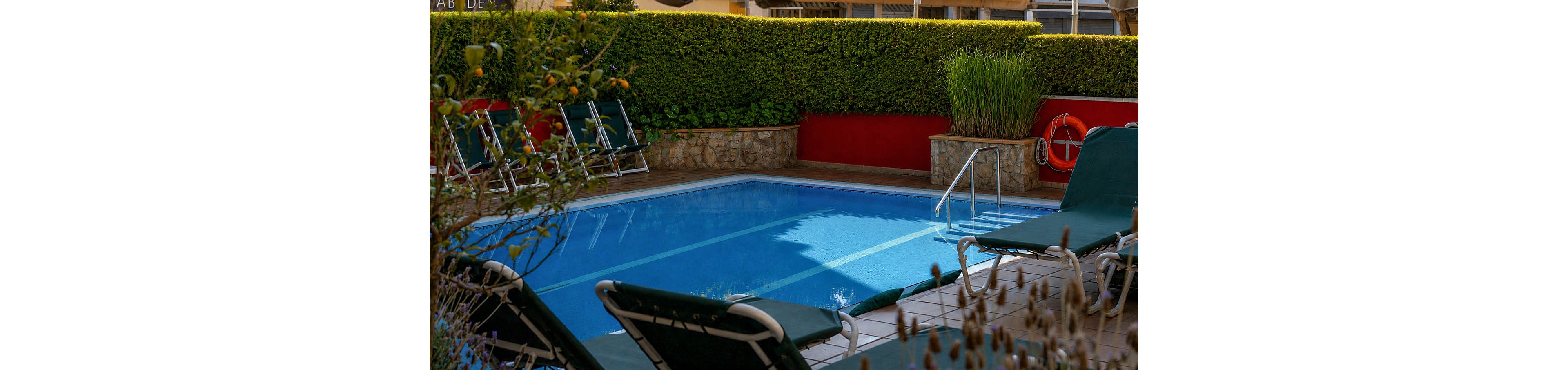 An outdoor swimming pool sits in a small courtyard surrounded by hedges and stone walls. Several green lounge chairs line the tiled poolside area, suggesting a relaxing place to sunbathe. A metal handrail leads into the clear blue water, and a life preserver hangs on the far wall for safety. The scene appears calm and inviting, with no people present.
